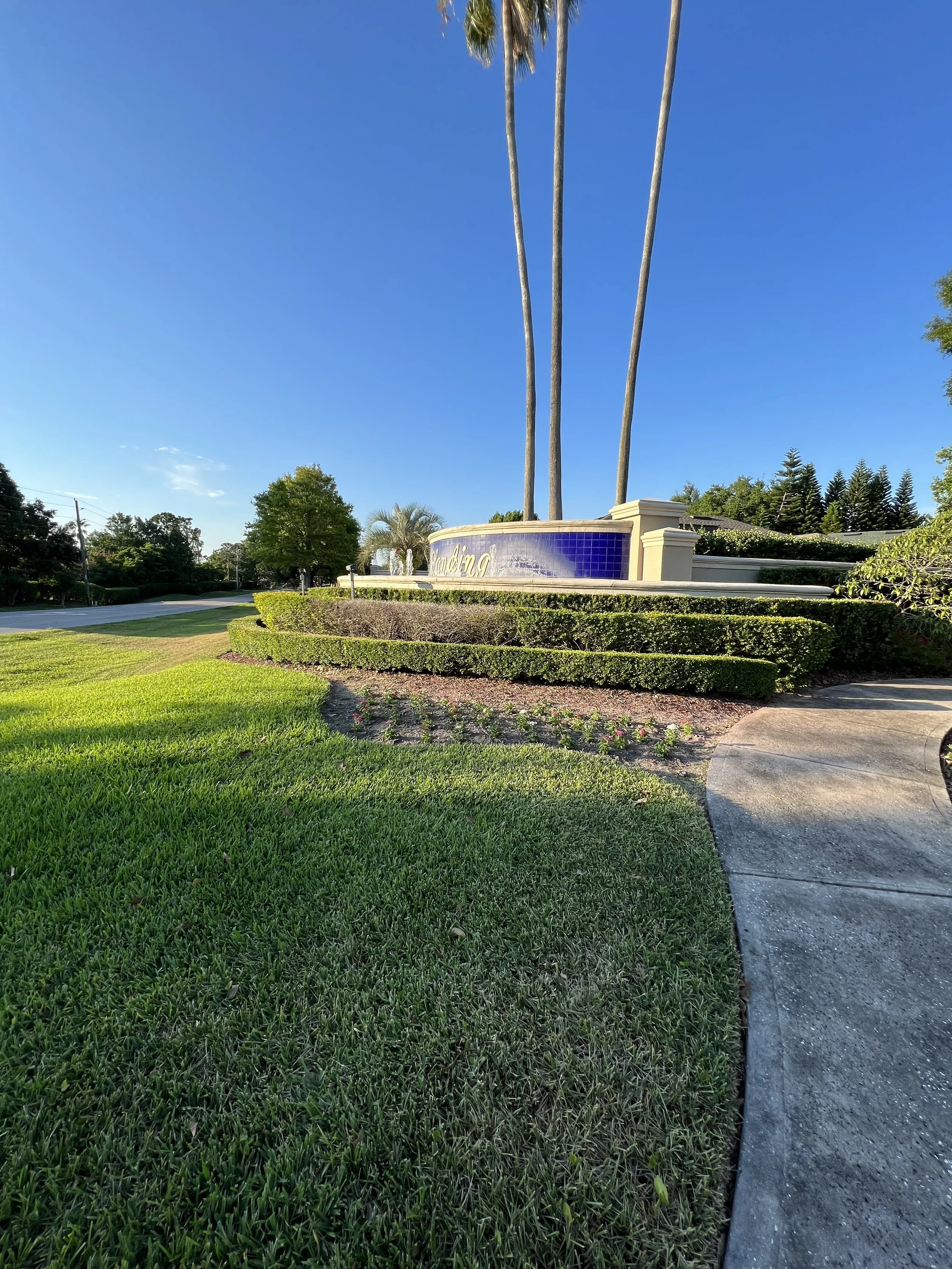 A landscaped area with green grass, a pathway, and a landscaped sign with palm trees and a blue sky in the background.