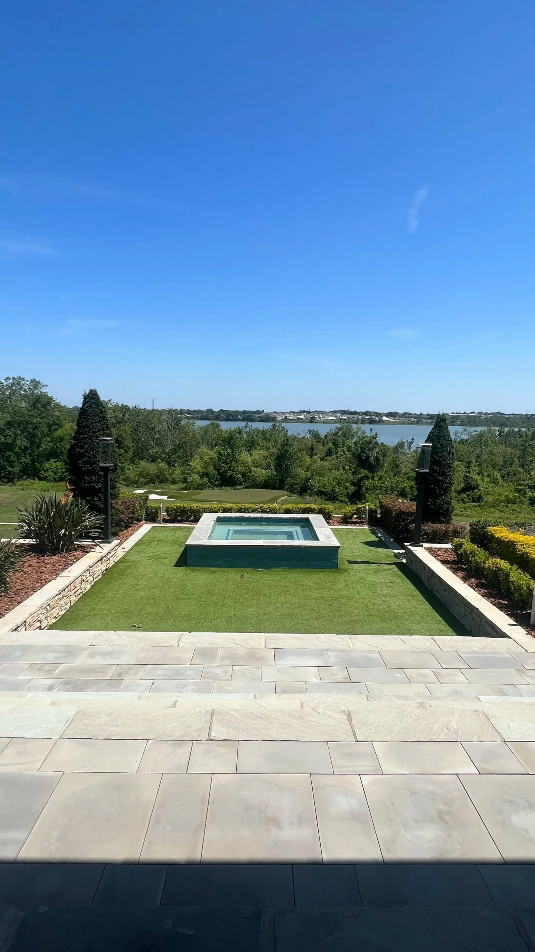 View from a patio with large beige tile flooring, a small rectangular pool or hot tub with a raised edge, surrounded by a well-manicured lawn, plants, and bushes, overlooking a body of water with trees and a distant shoreline under a bright blue sky.