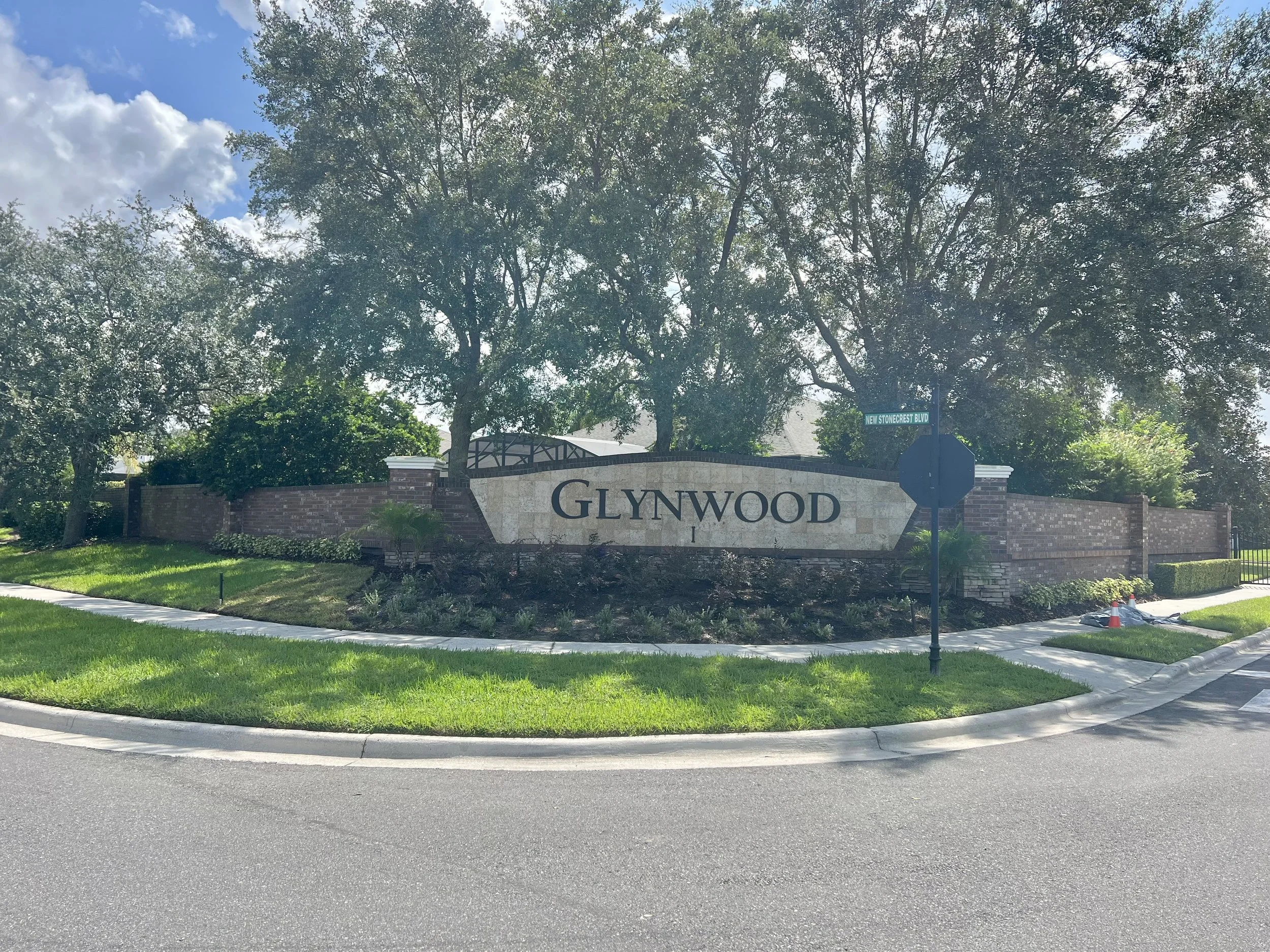 Entrance sign reading 'Glynnwood' surrounded by greenery, trees, and a sidewalk in front, with a street intersection sign indicating 'New Stonecrest Blvd' in an urban area.