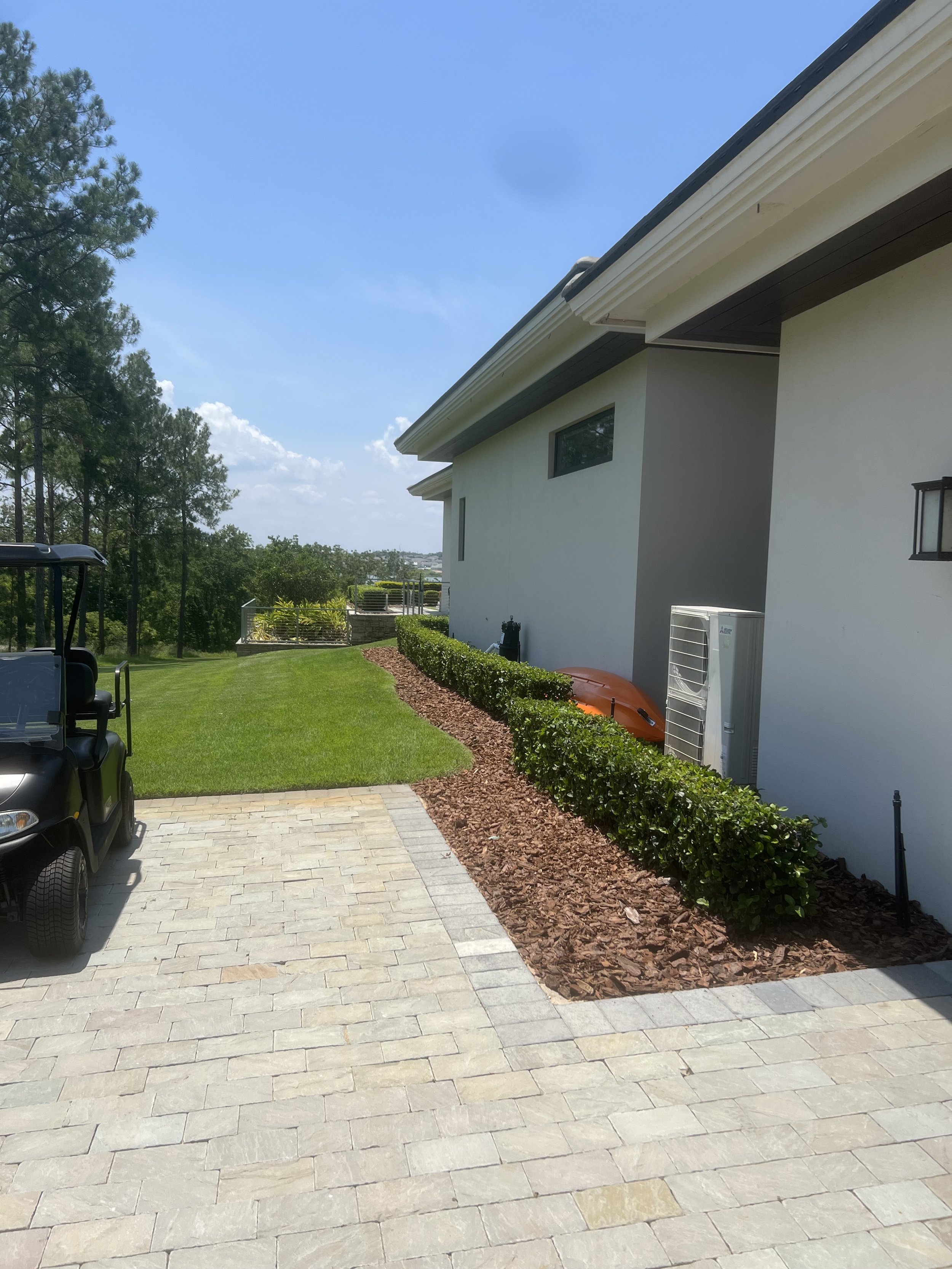 The image shows a modern backyard patio area with a paved surface, a house wall with a window, a bush border, and a lush green lawn. There is a golf cart parked on the patio. In the background, there are tall trees and a clear blue sky with some clou
