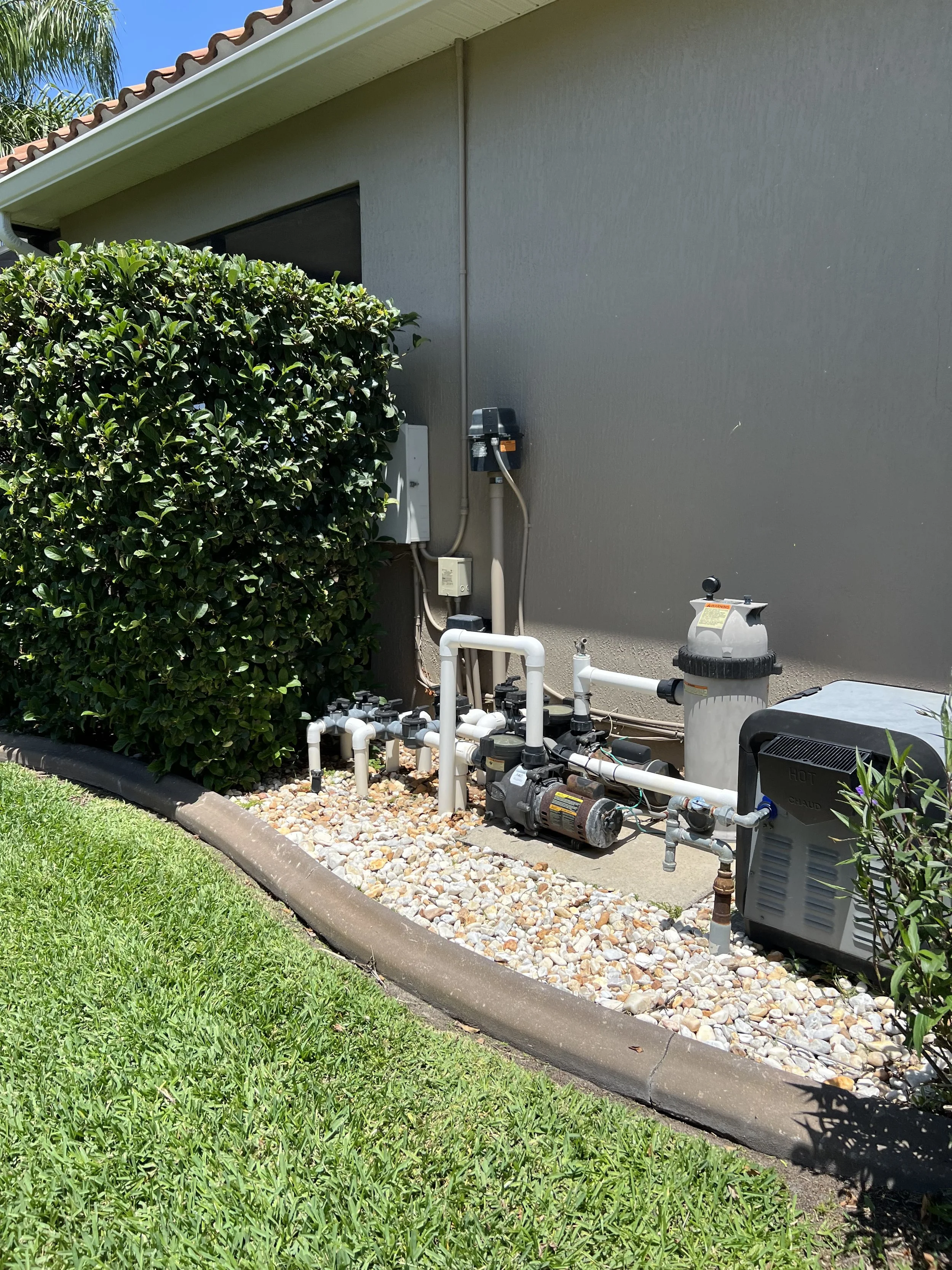Side view of a house exterior with a large hedge, white pipes, pump system, and pool equipment on a concrete slab, adjacent to a grassy lawn.