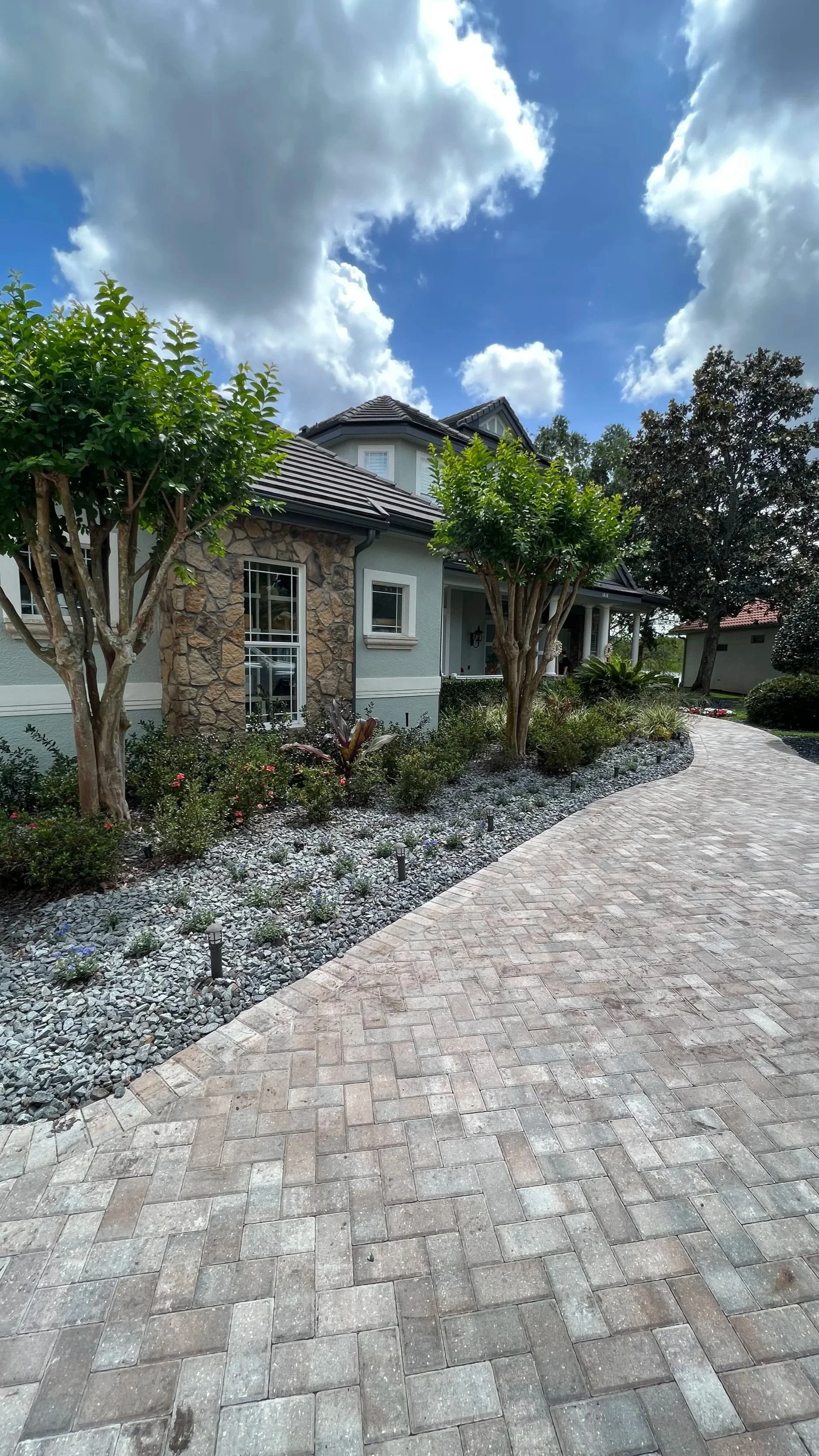 A brick driveway leading up to a house with a stone and stucco exterior, surrounded by trees and landscaped garden beds, under a partly cloudy sky.