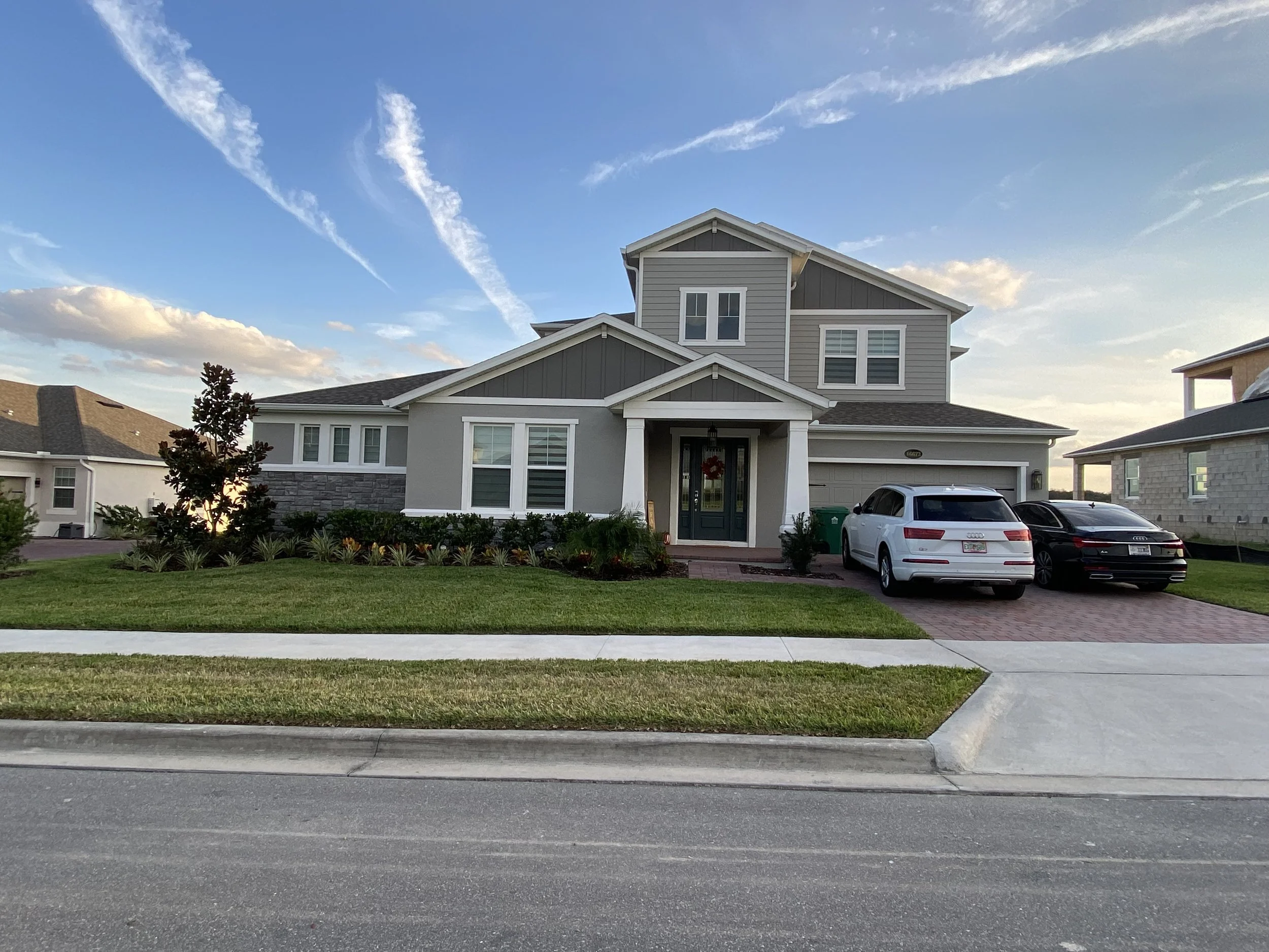 A modern two-story house with a gray roof, white and gray exterior, front porch, and a lawn with bushes. There are three cars parked in the driveway and a sidewalk in front. The sky is blue with some clouds and contrails.