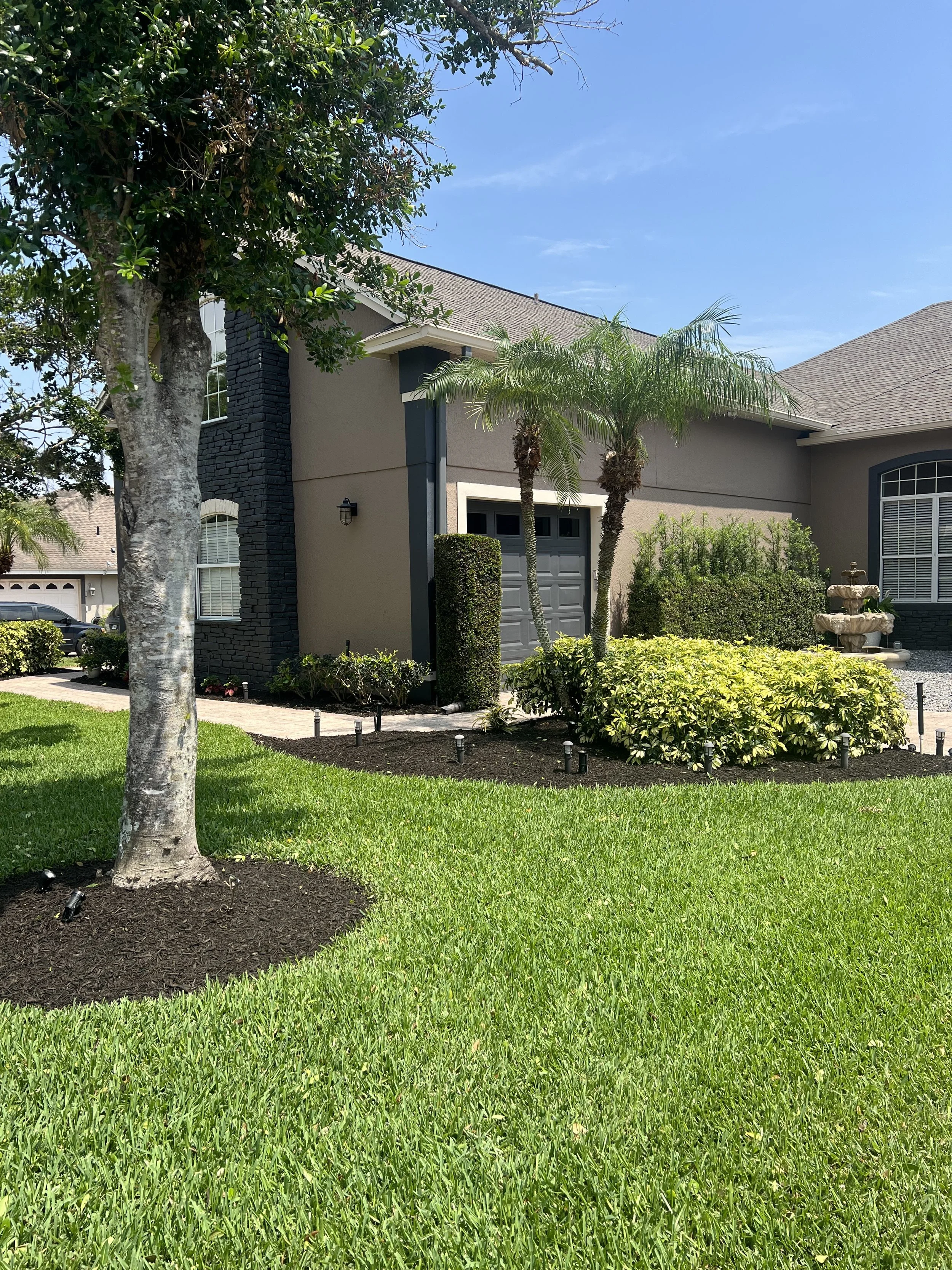 Front yard of a suburban house with green grass, trees, shrubs, and a fountain near the right side of the house, under a clear blue sky.