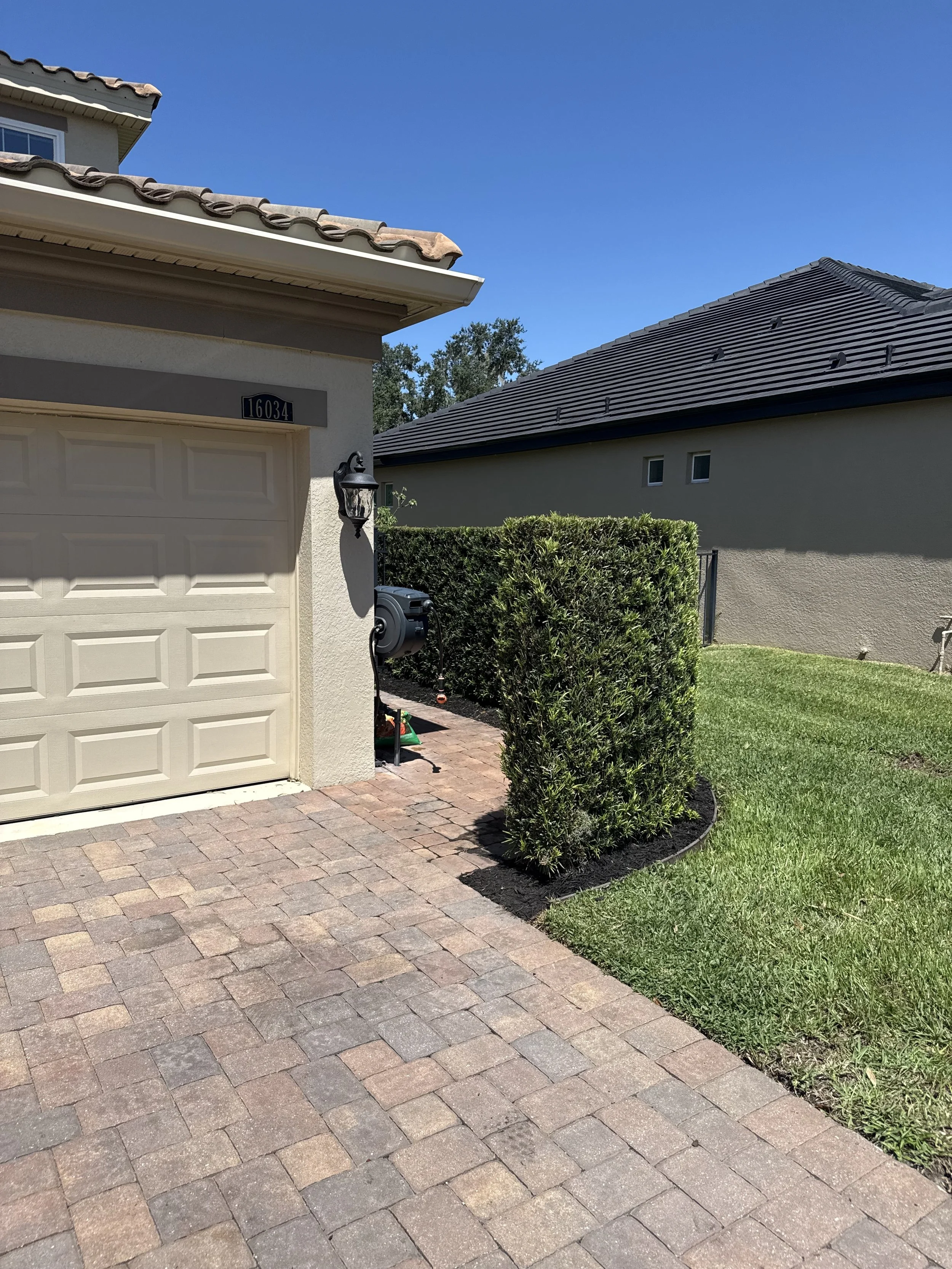 Front of a suburban house with beige garage door, address number 16034, porch light, greenery, brick driveway, neighboring house with dark roof, and blue sky.