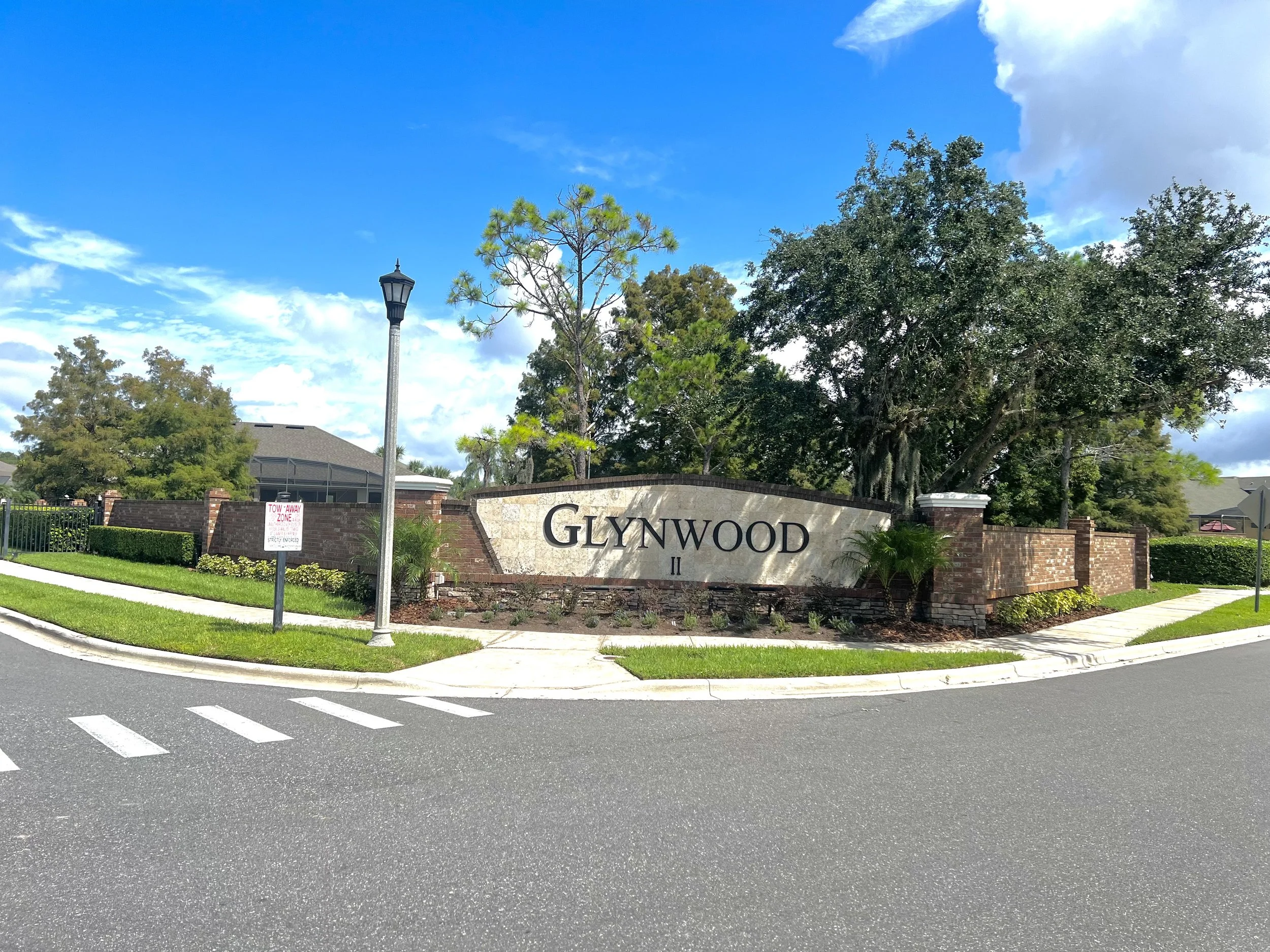 Street view of Glynwood community entrance with a stone sign, trees, a lamppost, a brick fence, and a crosswalk in the foreground.
