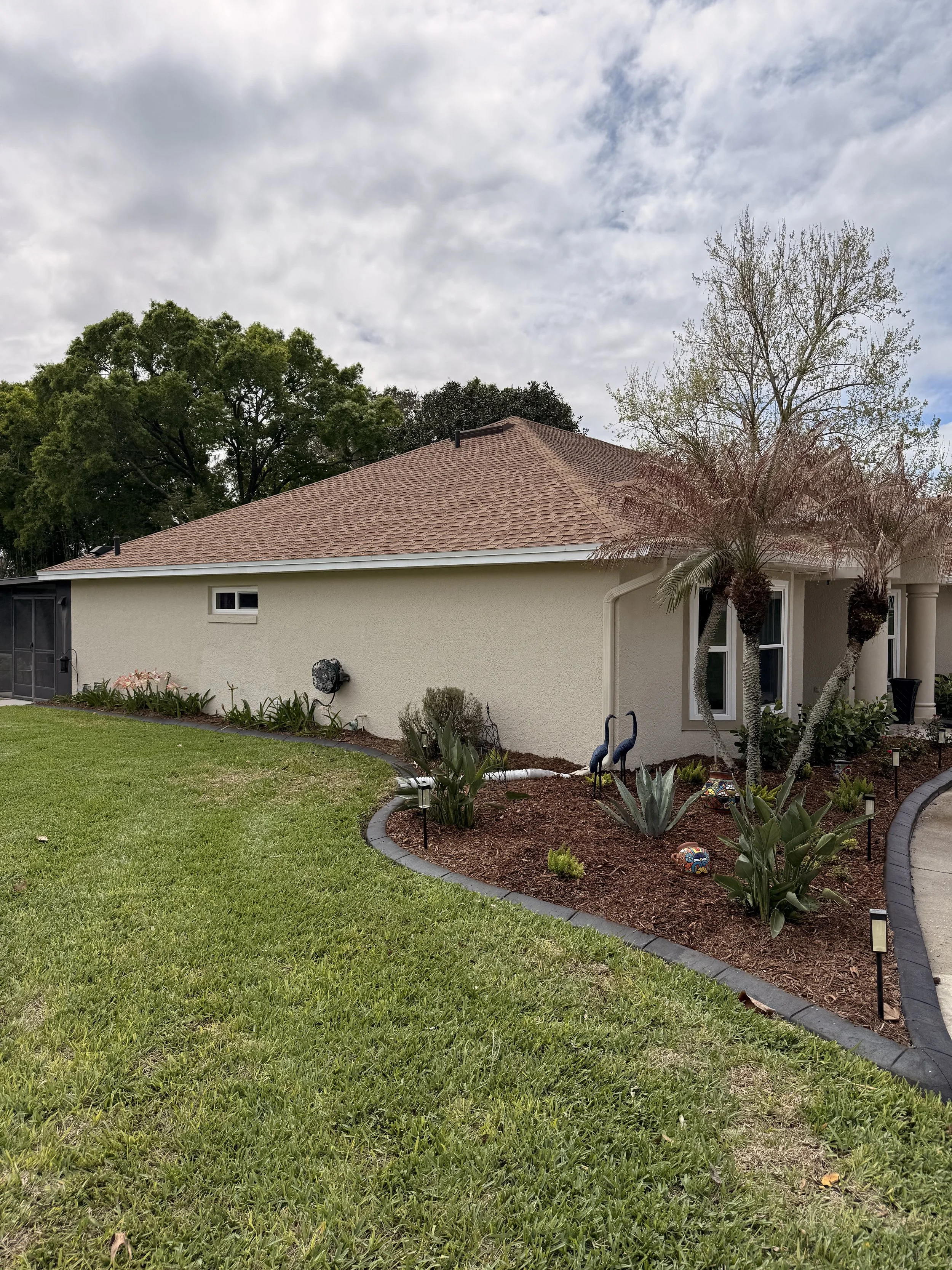 A backyard with a well-maintained green lawn, garden bed with plants and decorative statues, and a house with beige exterior walls, a sloped roof, and a small window. Tall trees are in the background under a partly cloudy sky.