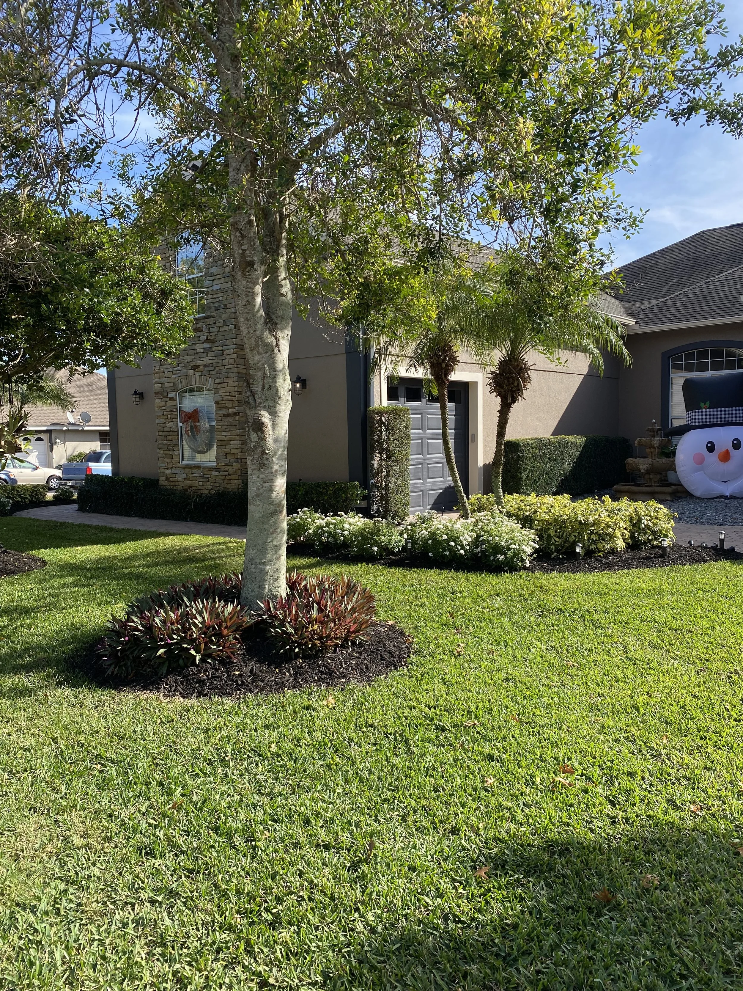 View of a residential front yard with green grass, small shrubs, trees, and a house with a stone and stucco exterior, garage door, and holiday decorations on the window, along with a snowman decoration and pathway.