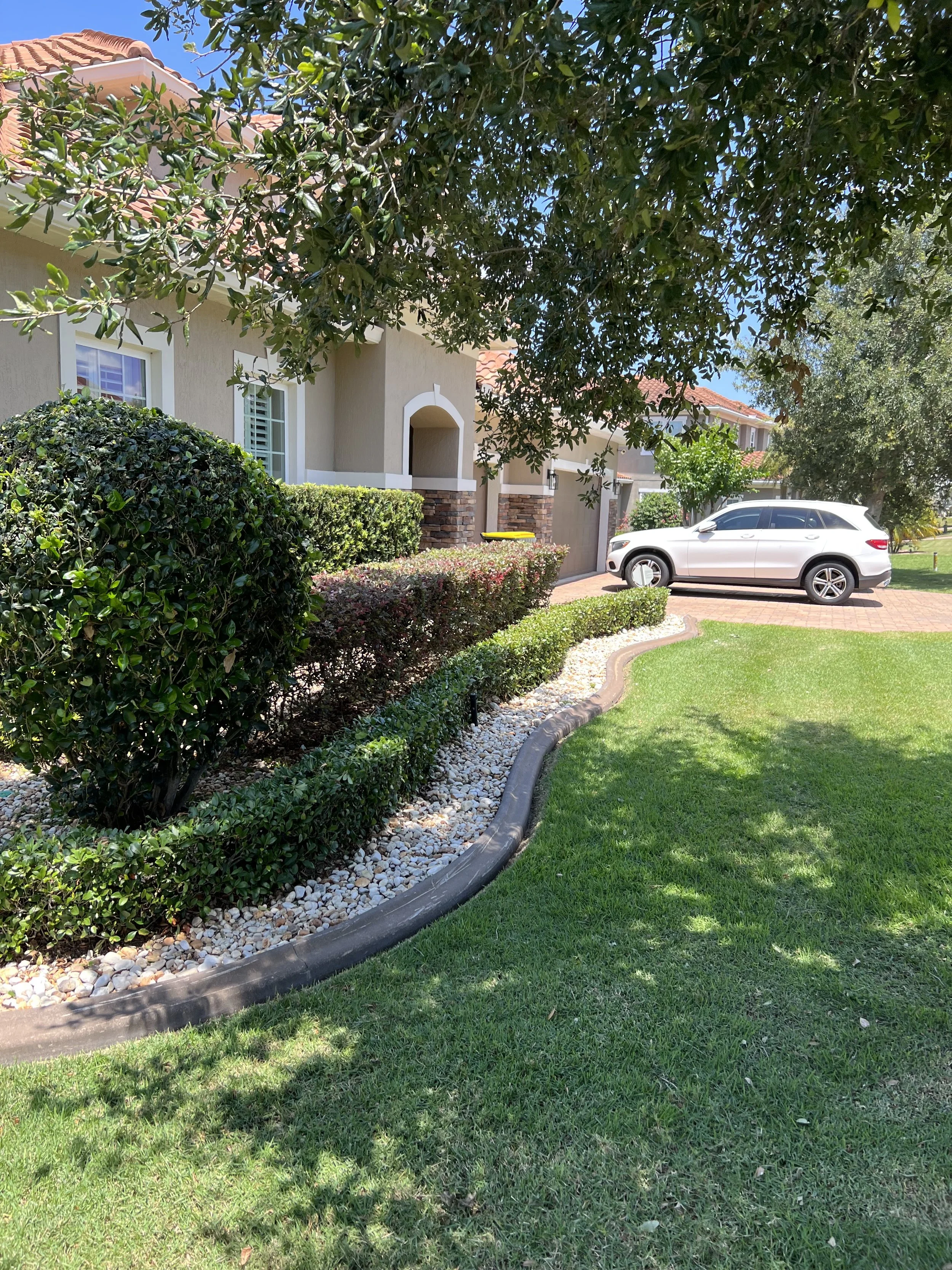 A residential house with a beige exterior, white trim, and a red tile roof. There are green bushes and trees in front, along with a curved garden bed lined with white rocks. A white SUV is parked near the house, and shadows from the trees cover part 