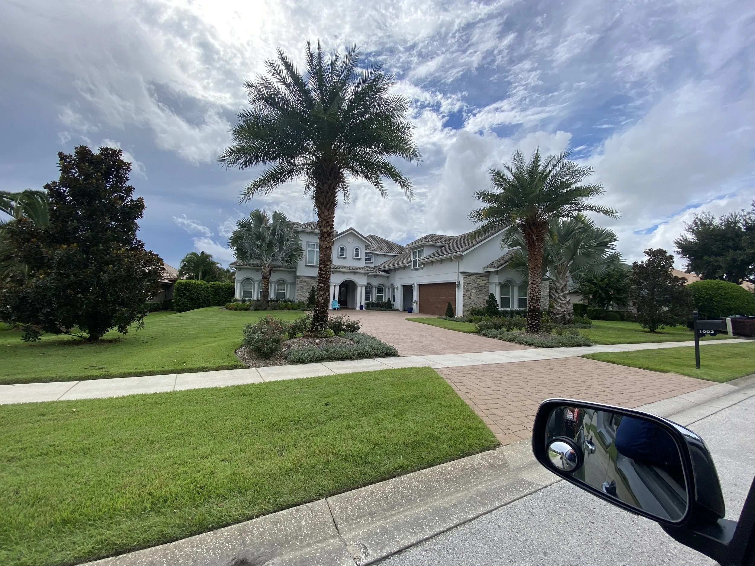 Front yard of a large house with a lawn, palm trees, shrubs, and a driveway. The house has a white exterior with stone accents and a tiled roof. The sky is partly cloudy.
