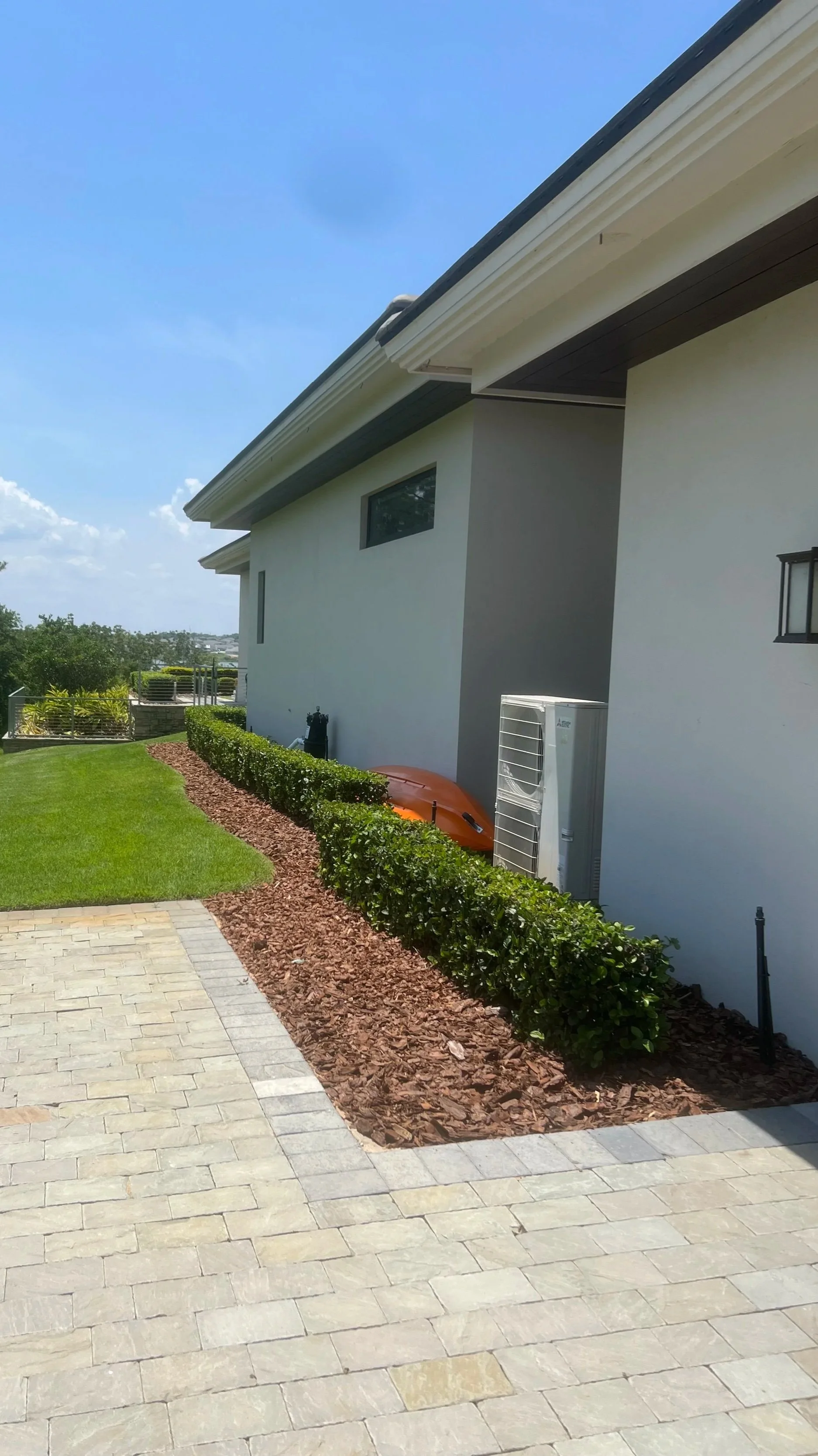 Backyard with green lawn, bushes, mulch, and paved patio, with a house wall and roof, an air conditioning unit, a kayak, and a water hose.