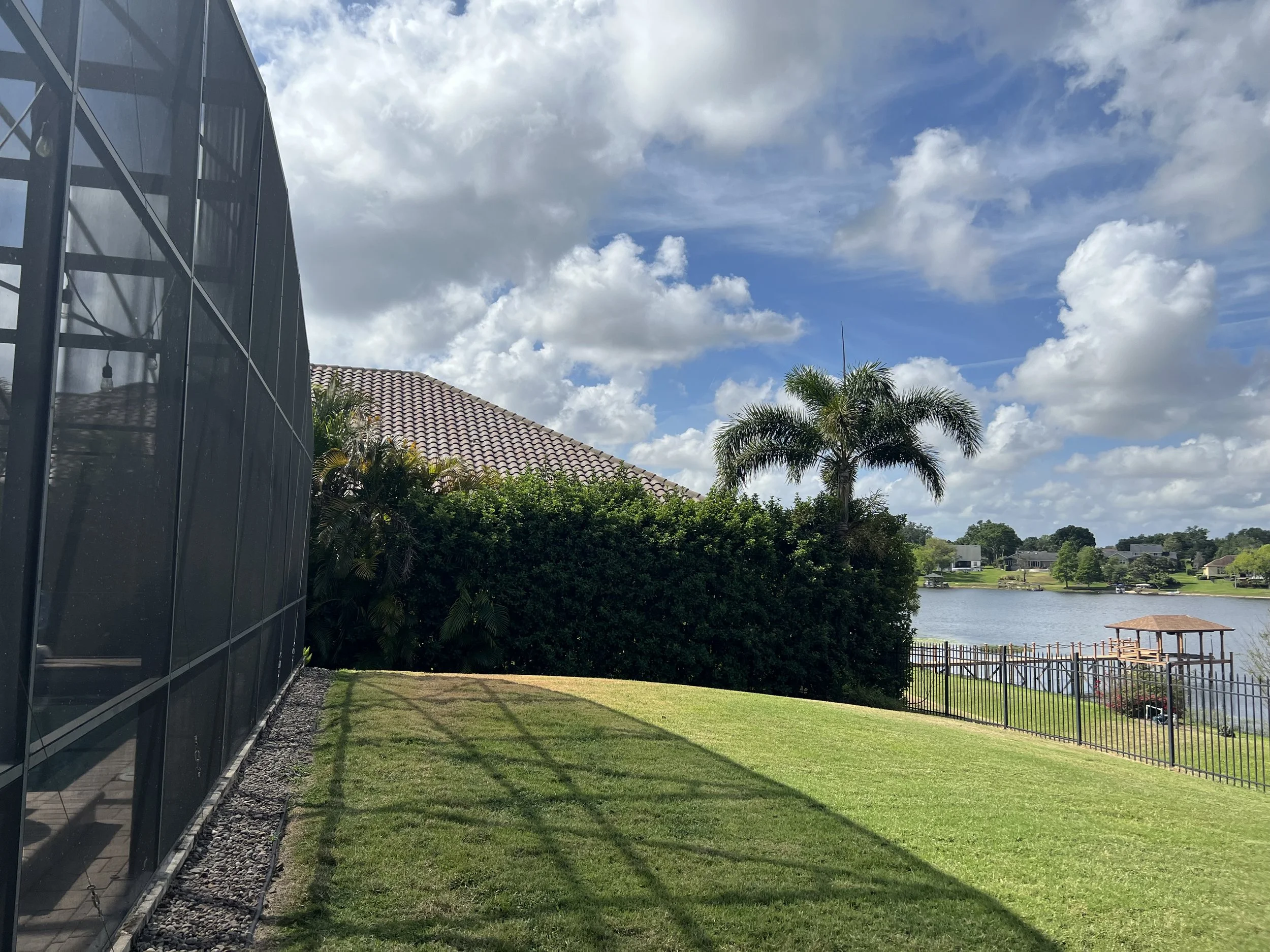 View of a backyard with green grass, a pond, and a black fence. On the left side, there's a modern building with dark reflective glass walls. In the background, there are trees, shrubs, a house with a tiled roof, and a partly cloudy sky with white and gray clouds.