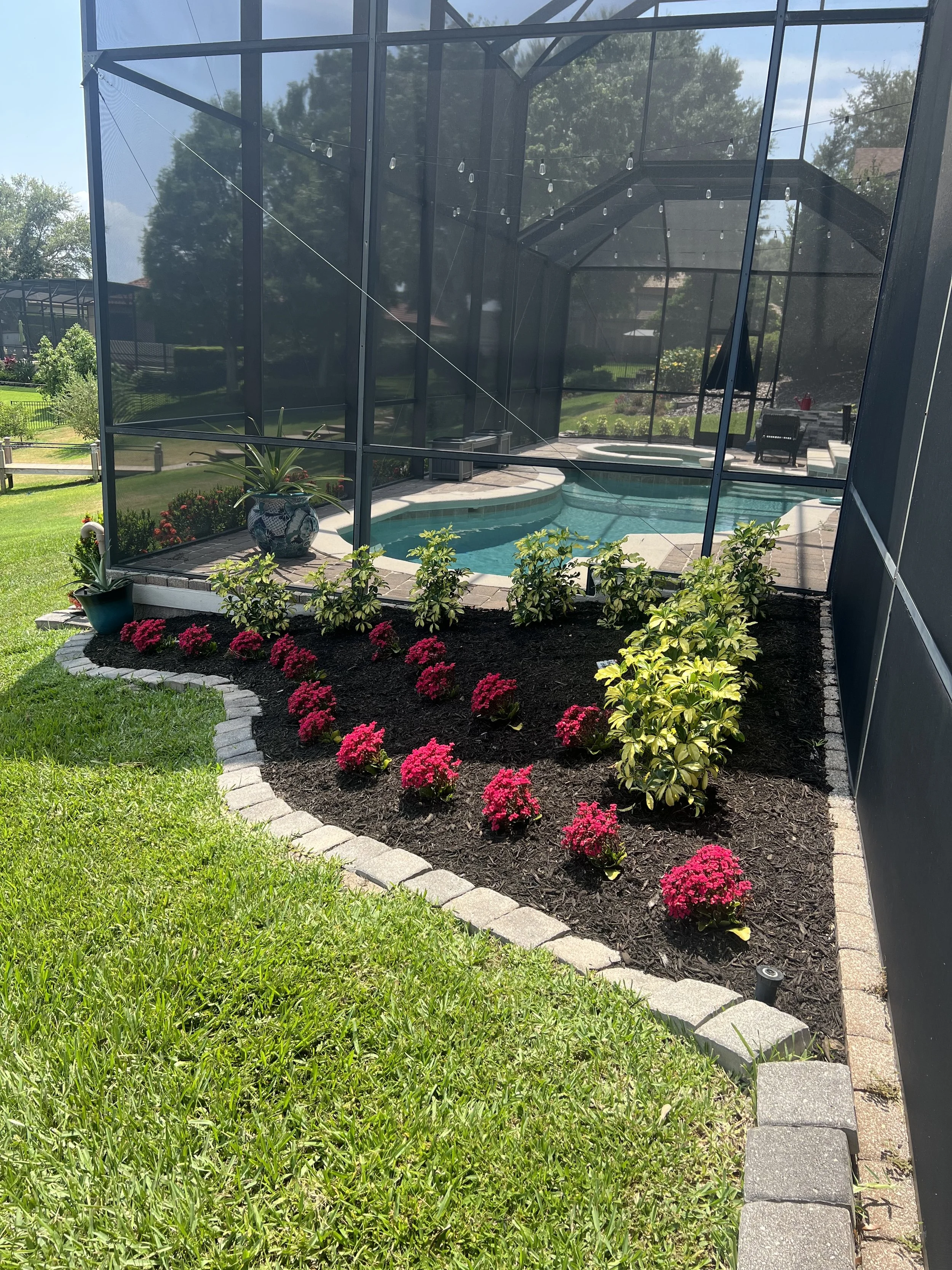 A backyard with a screened-in pool area, potted plants, pink flowers, and a garden bed with various plants and shrubs.