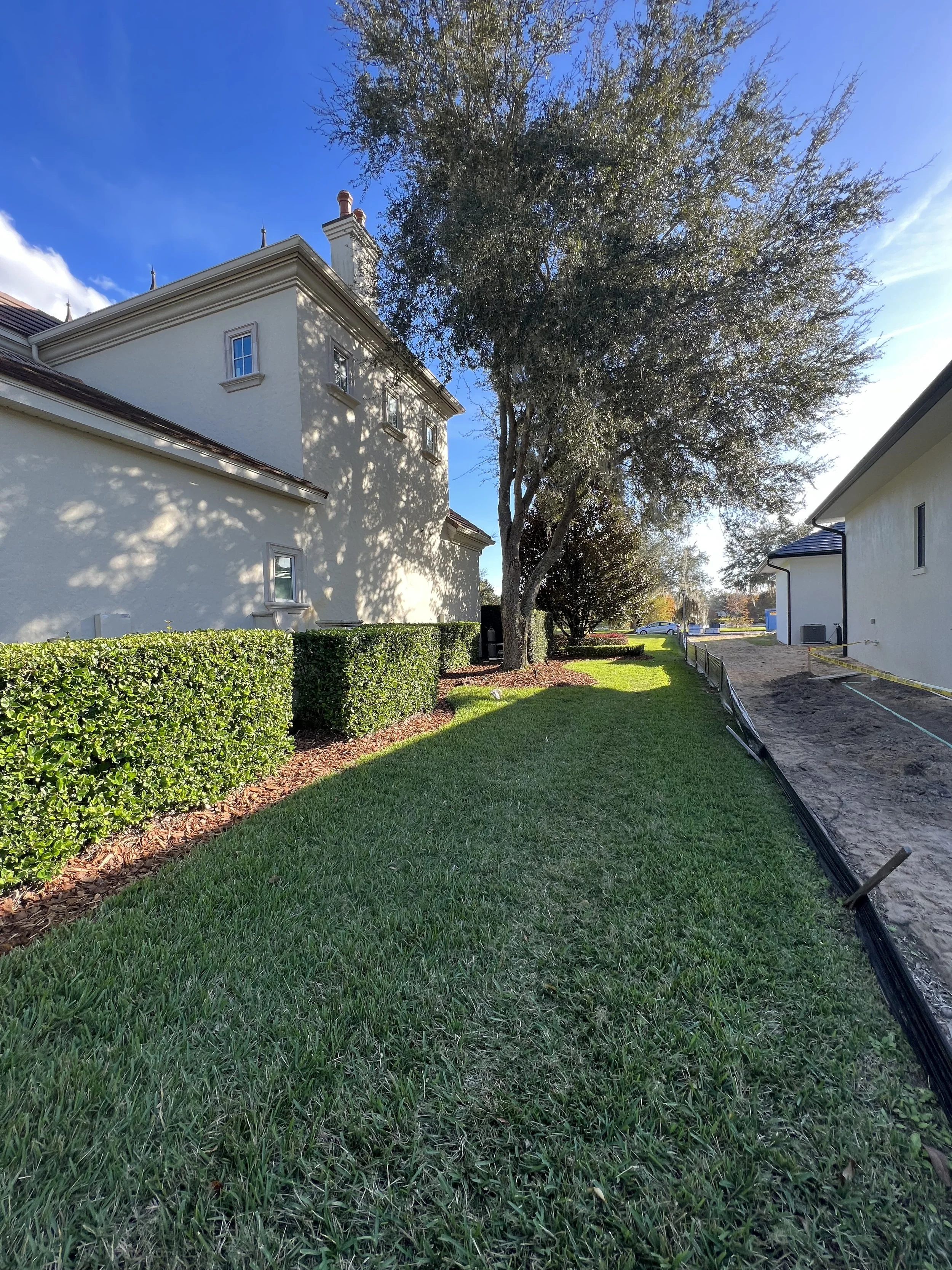 A suburban neighborhood scene with a lush green lawn, well-trimmed hedge, tall trees, and a white house with a brown roof. There is a construction area with dirt and a black pipe running along the ground on the right side of the image. The sky is par