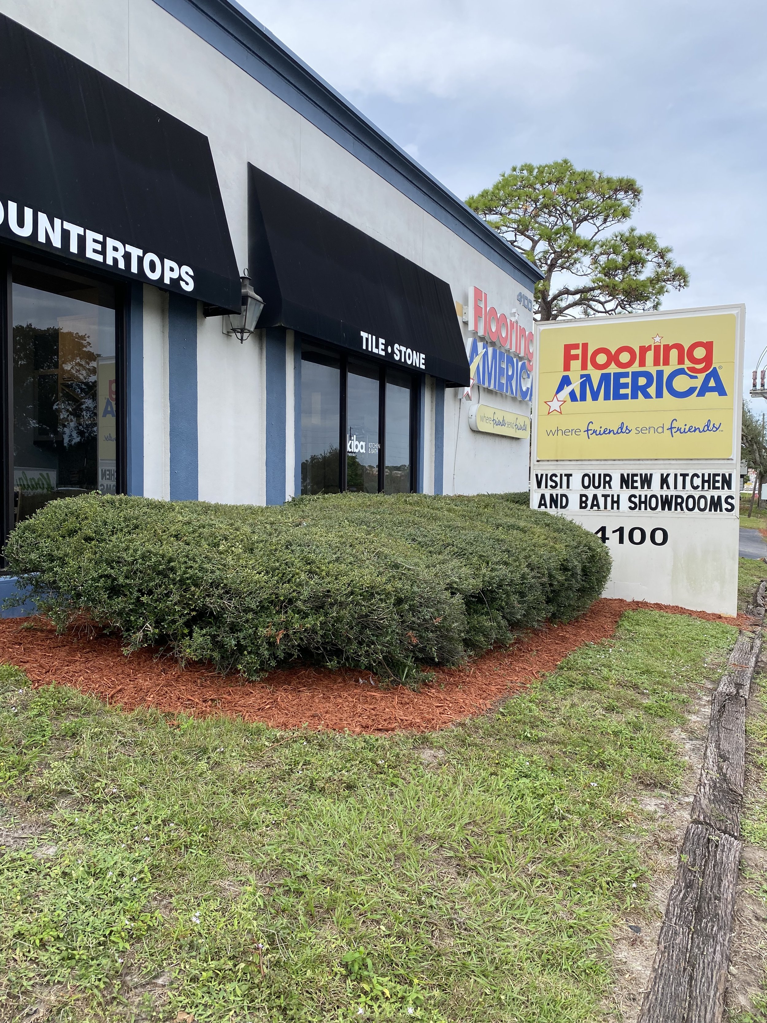 A storefront with black awnings, a sign for Flooring America, and landscaping with bushes and mulch in front.