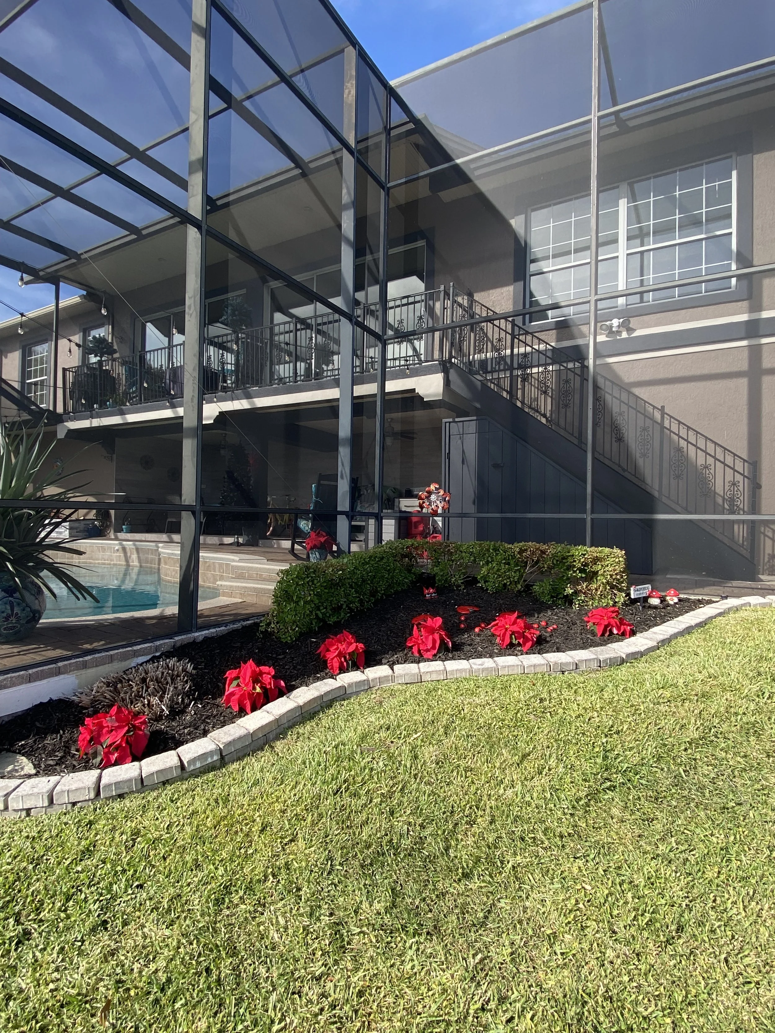 A modern residential house with a large glass enclosed patio area. There is a staircase leading to an upper deck with metal railing, and outdoor furniture. In the foreground, there is a well-maintained lawn with a flower bed containing red flowers an