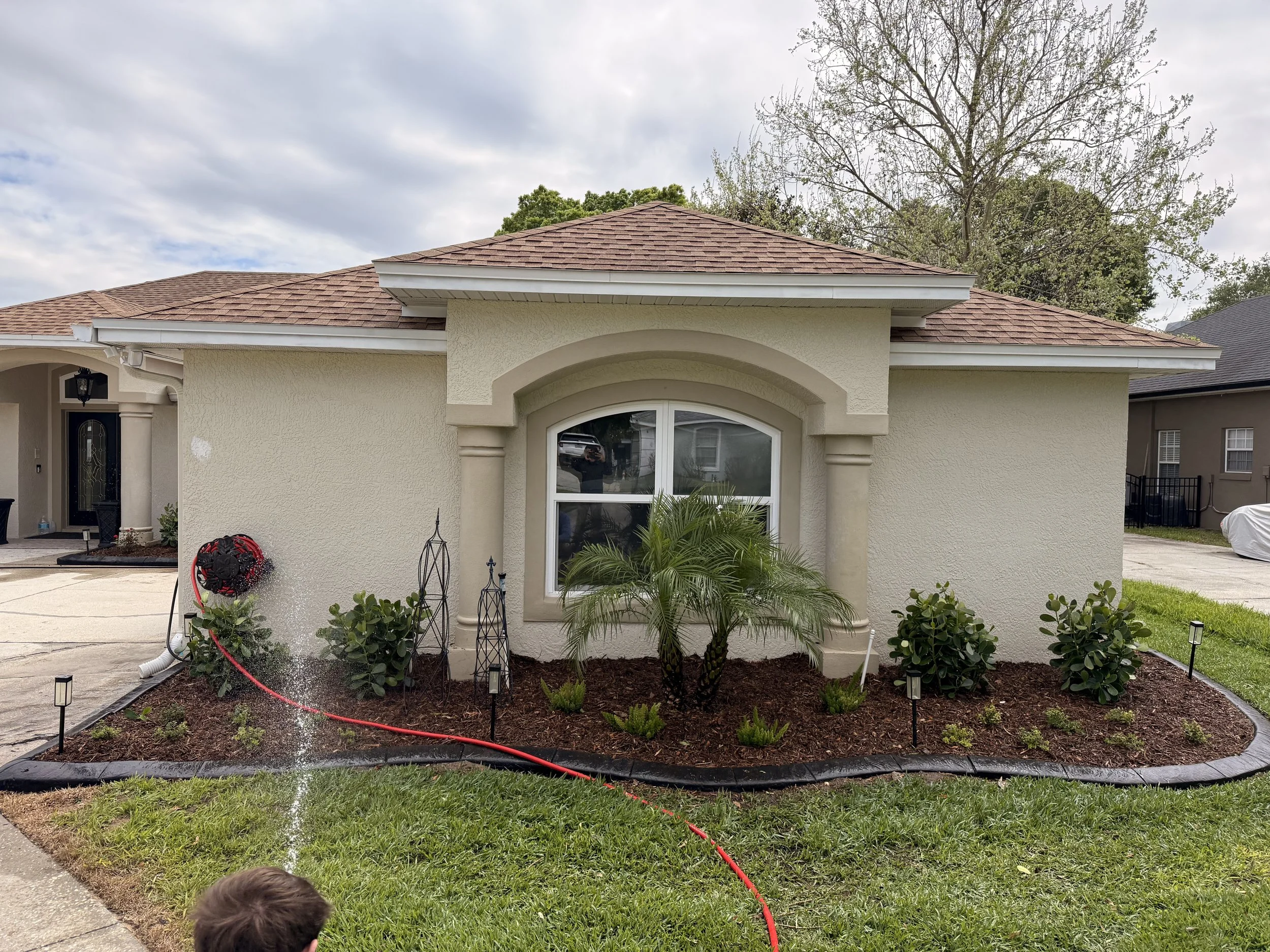 The front of a house with beige stucco walls and a brown shingle roof, surrounded by a small garden with plants and lawn. A person is partially visible at the bottom of the image, and the sky is cloudy.