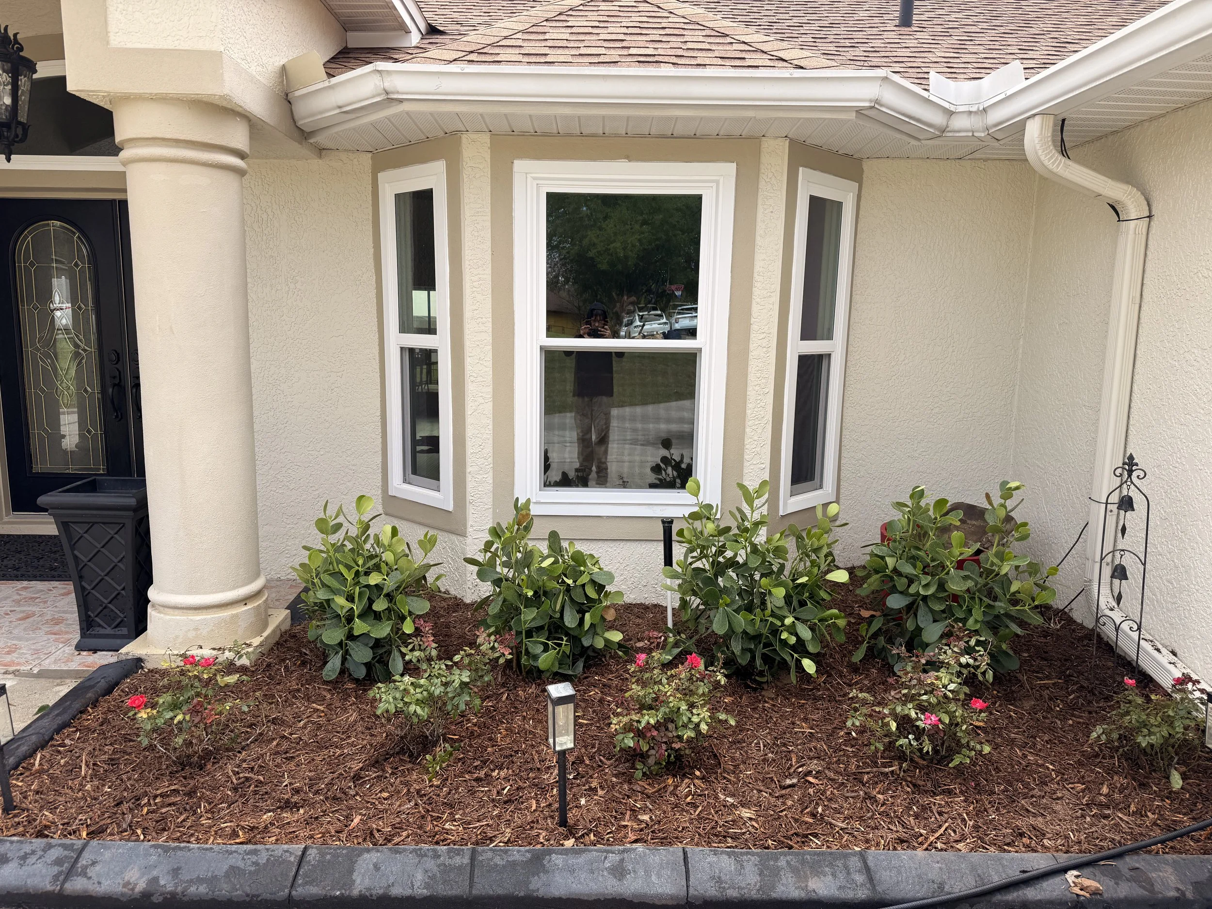 Front porch with a beige house featuring a large bay window, some plants and flowers in front, and a black door with decorative glass panel. The porch has a tiled floor and a column supporting the roof.