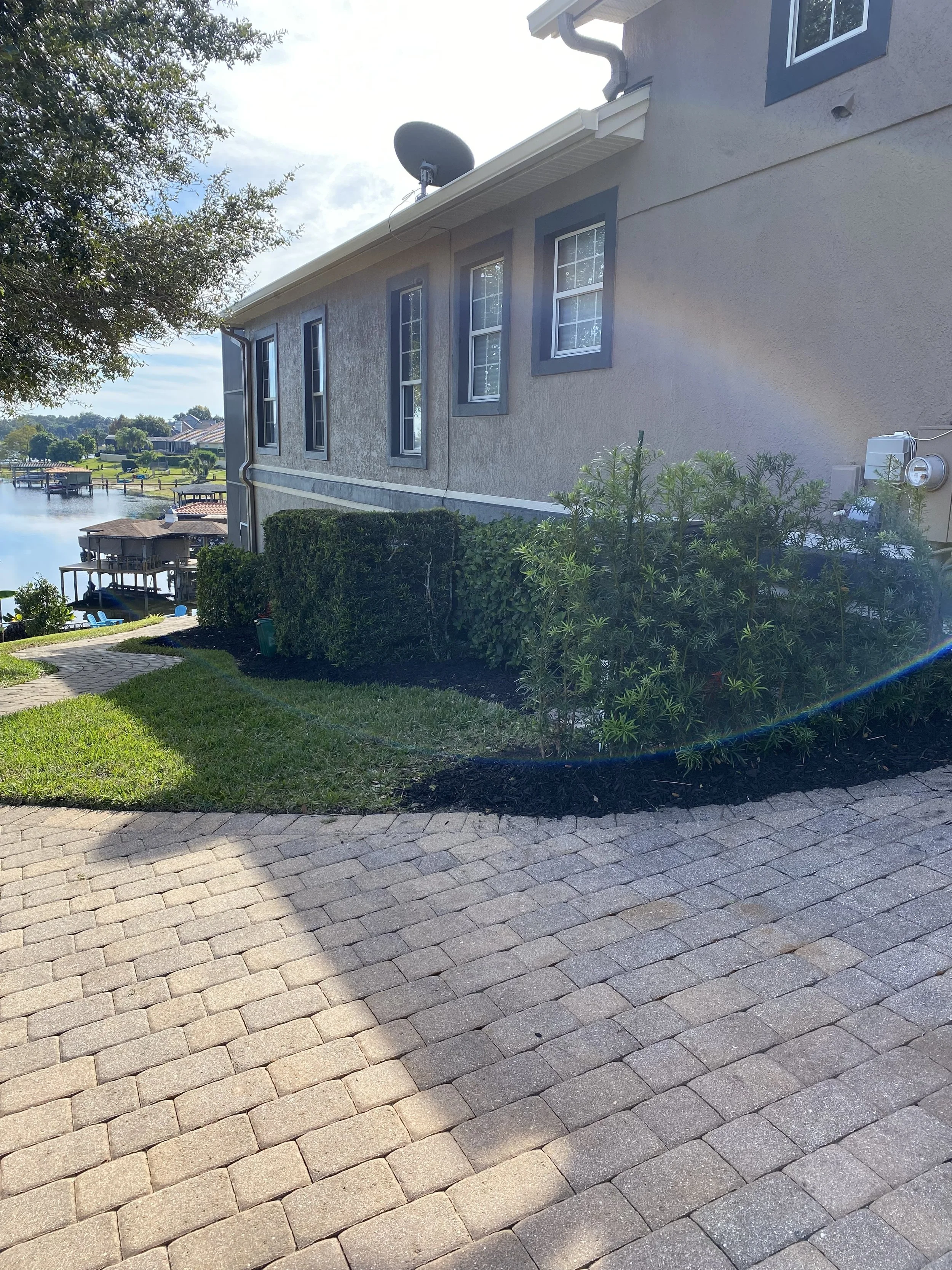 Backyard view with a paved stone patio, green bushes, a grassy area, and a house with multiple windows and a satellite dish on the roof.