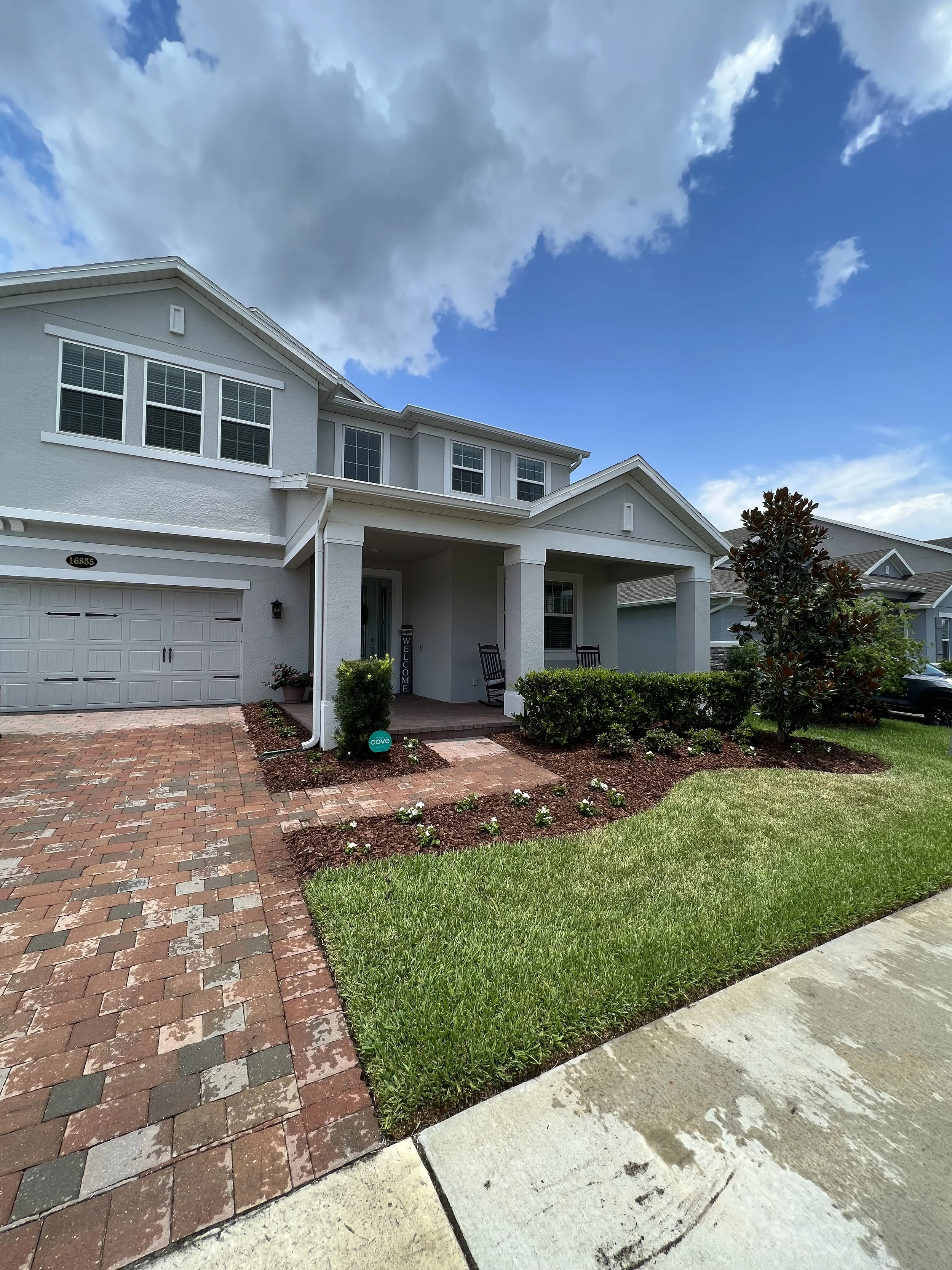 Front view of a white two-story house with a garage, a small porch with chairs, a landscaped yard with bushes and flowers, a brick walkway, and a sidewalk, under a partly cloudy sky.