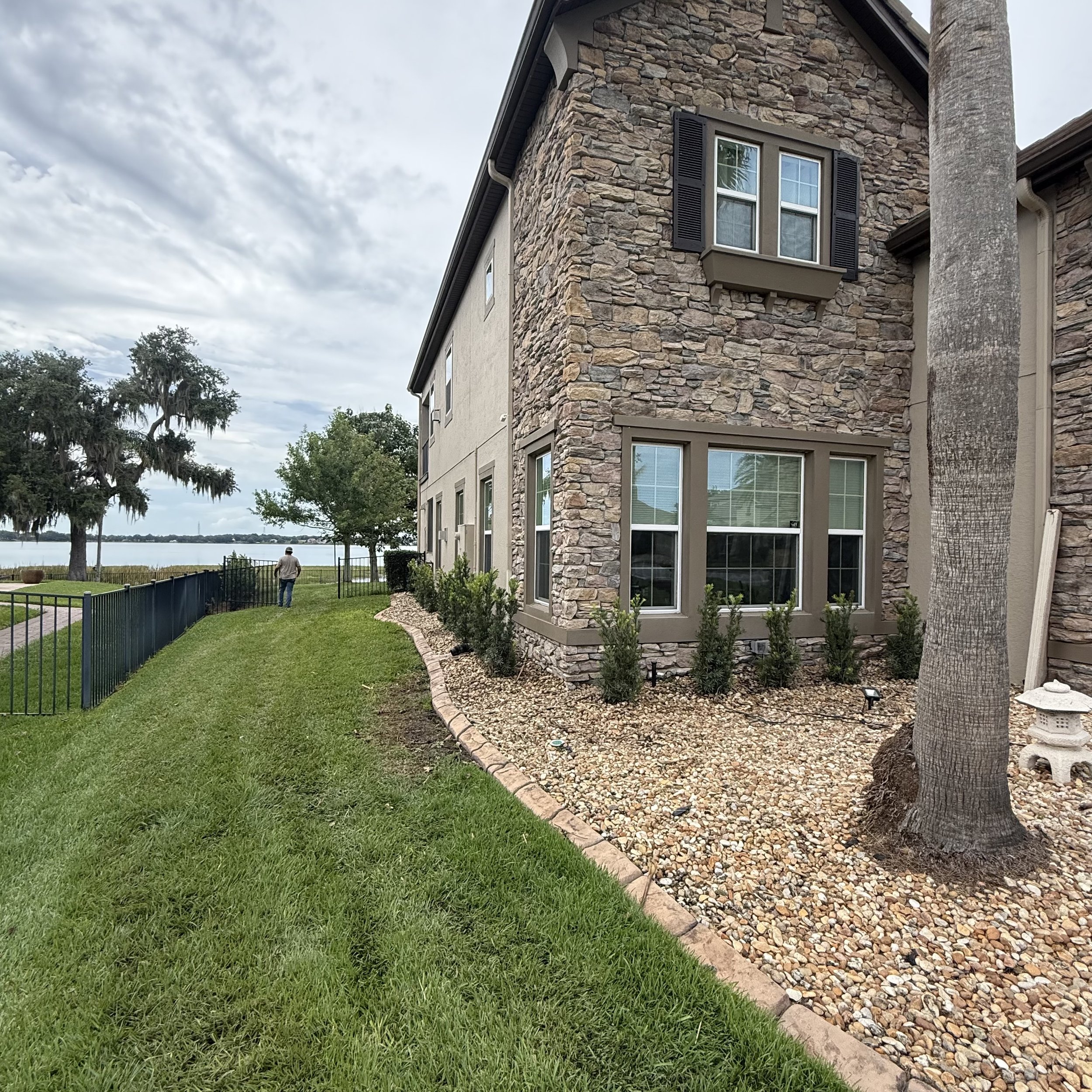 View of a residential building with stone and stucco exterior, multiple windows with black shutters, a grassy yard, and a person walking near the water in the background, under a cloudy sky.