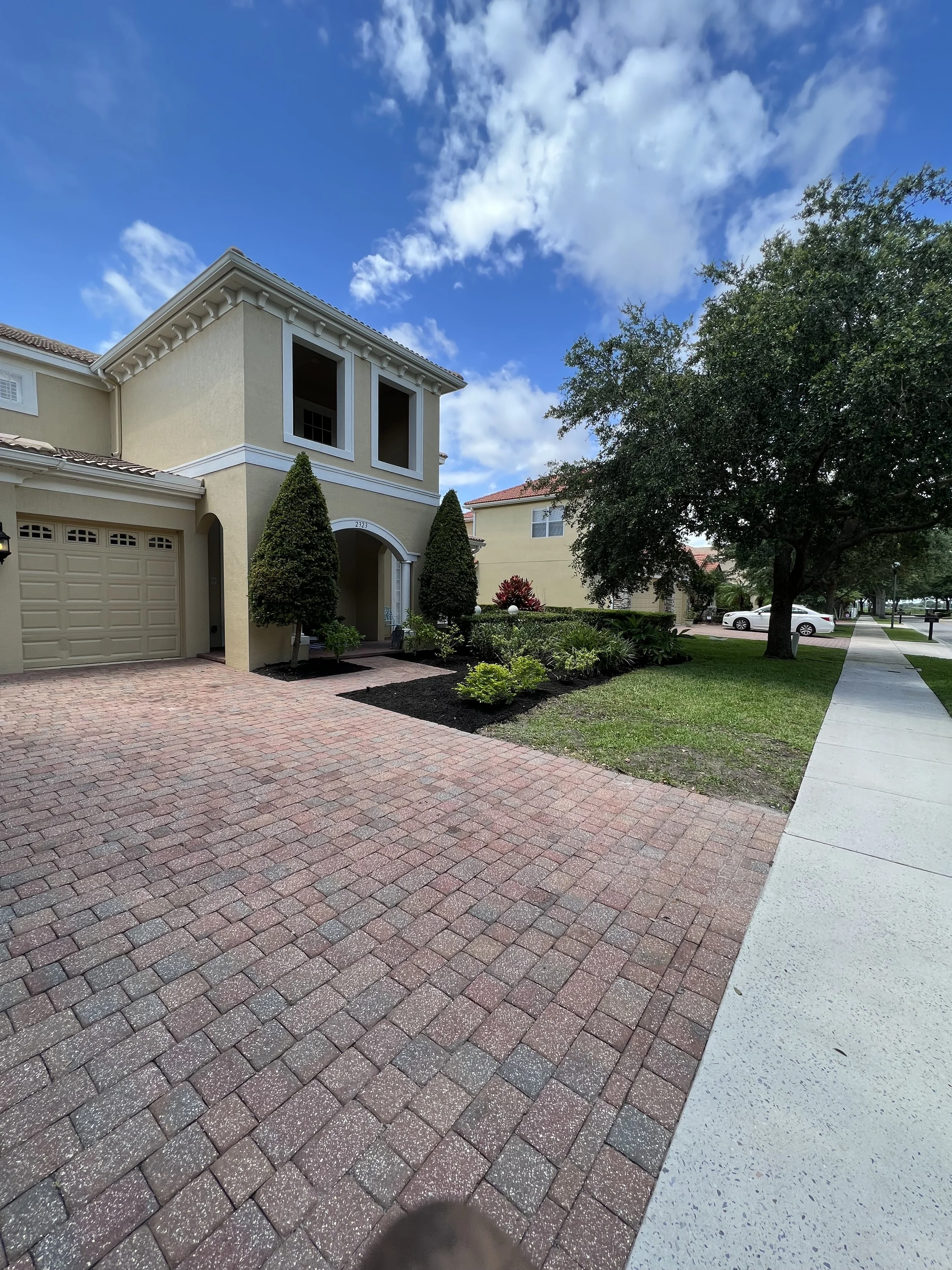 Residential neighborhood with houses, trees, a sidewalk, and a paved driveway under a partly cloudy sky.