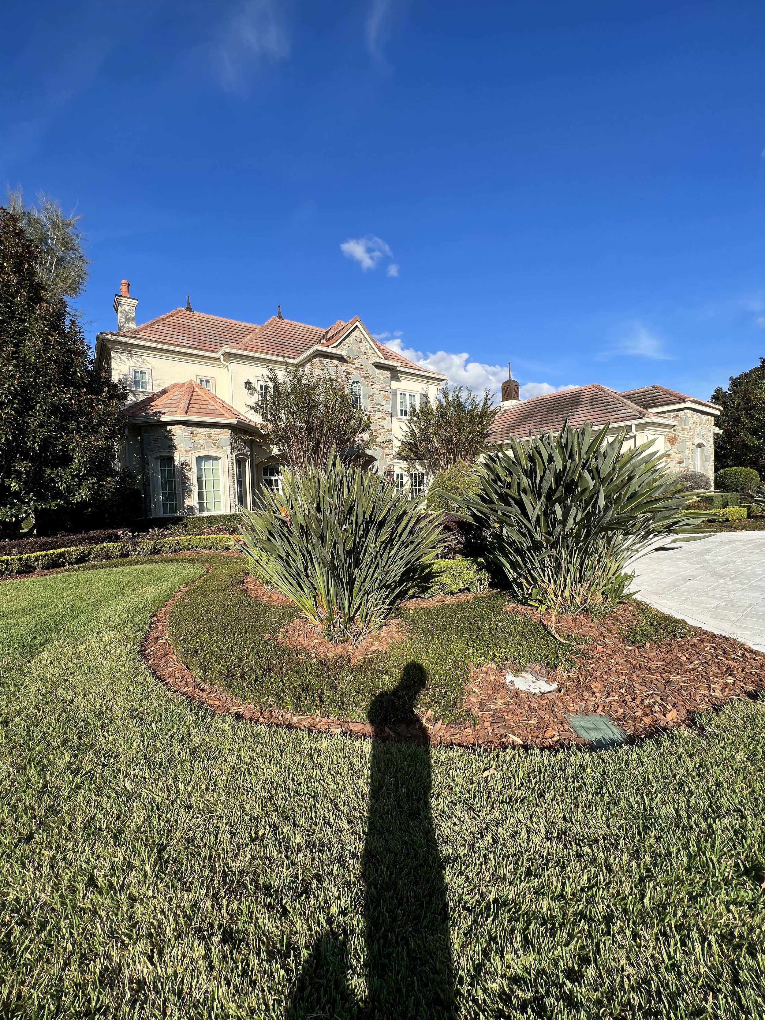 A large house with a stone facade and red tile roof, surrounded by landscaped green lawn, bushes, and trees, under a bright blue sky with a few small clouds.