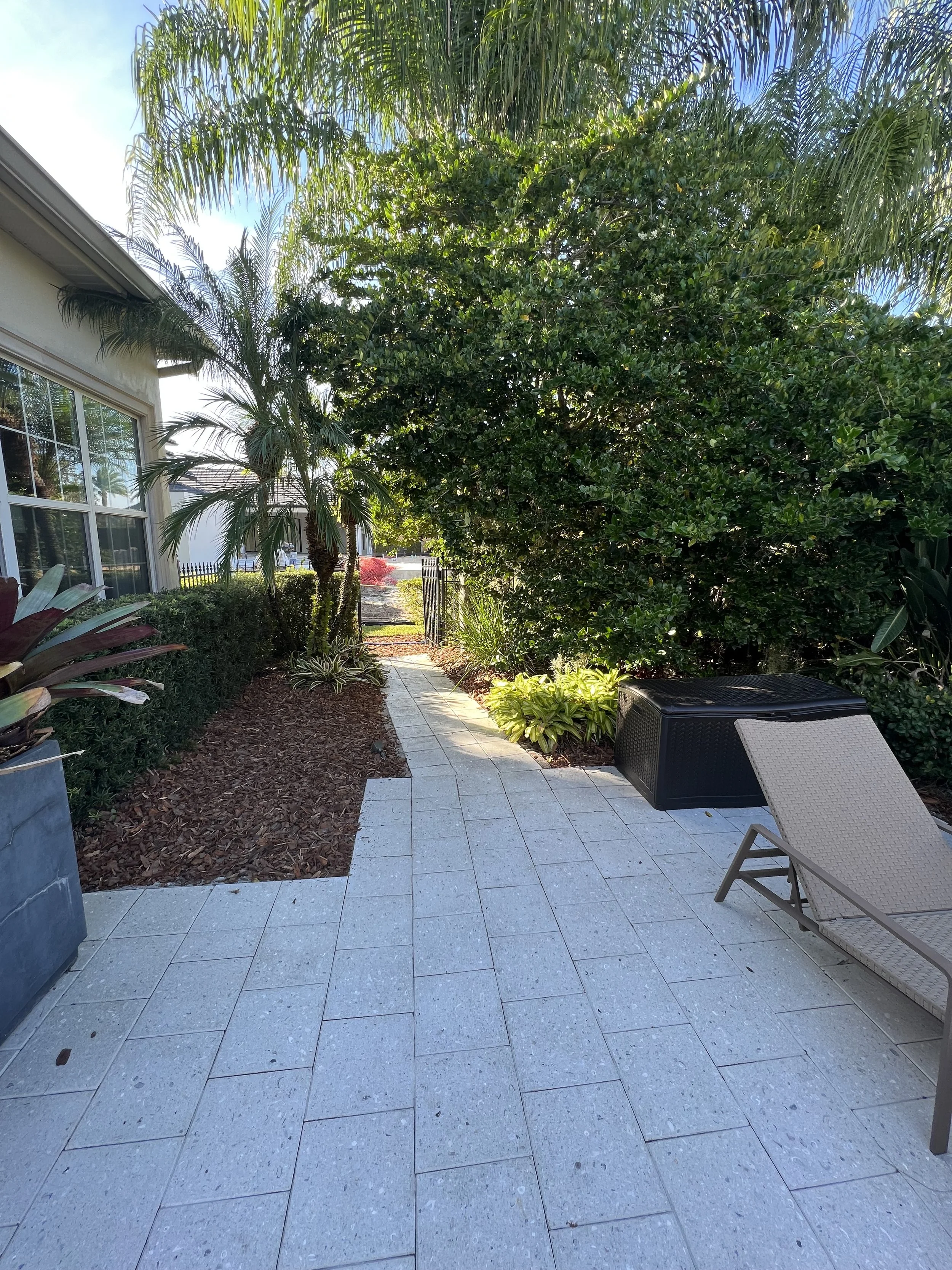 A backyard patio with light gray stone tiles, a lounge chair, lush green bushes and trees including palms, and a view of a neighboring house with a white exterior wall and large windows.