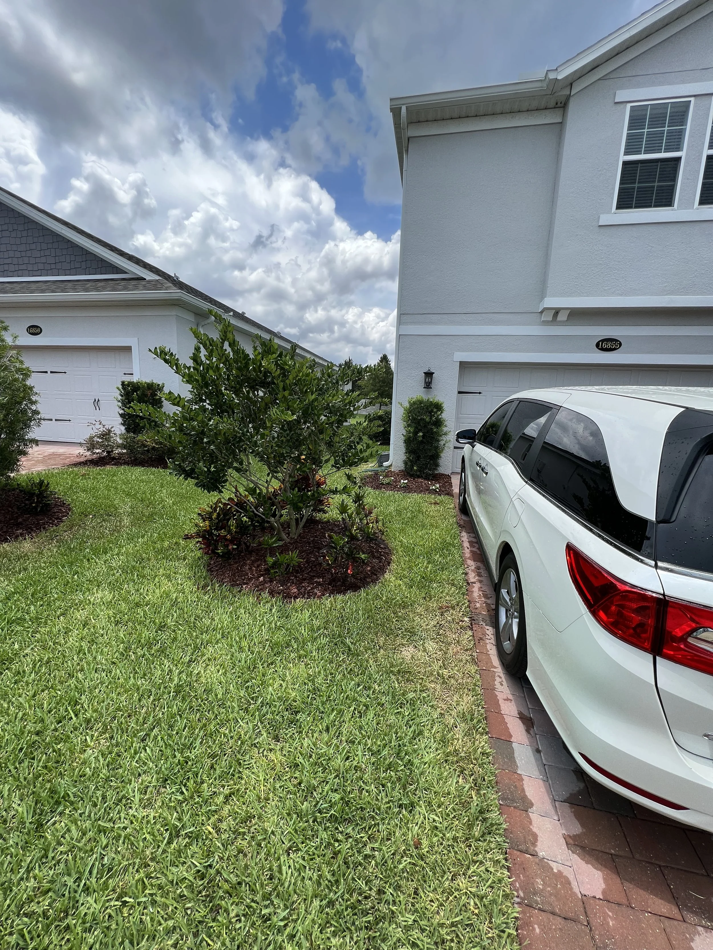 A residential scene showing a white car parked on a brick-lined driveway next to a white house with green shrubbery and a lawn, cloudy sky in the background.