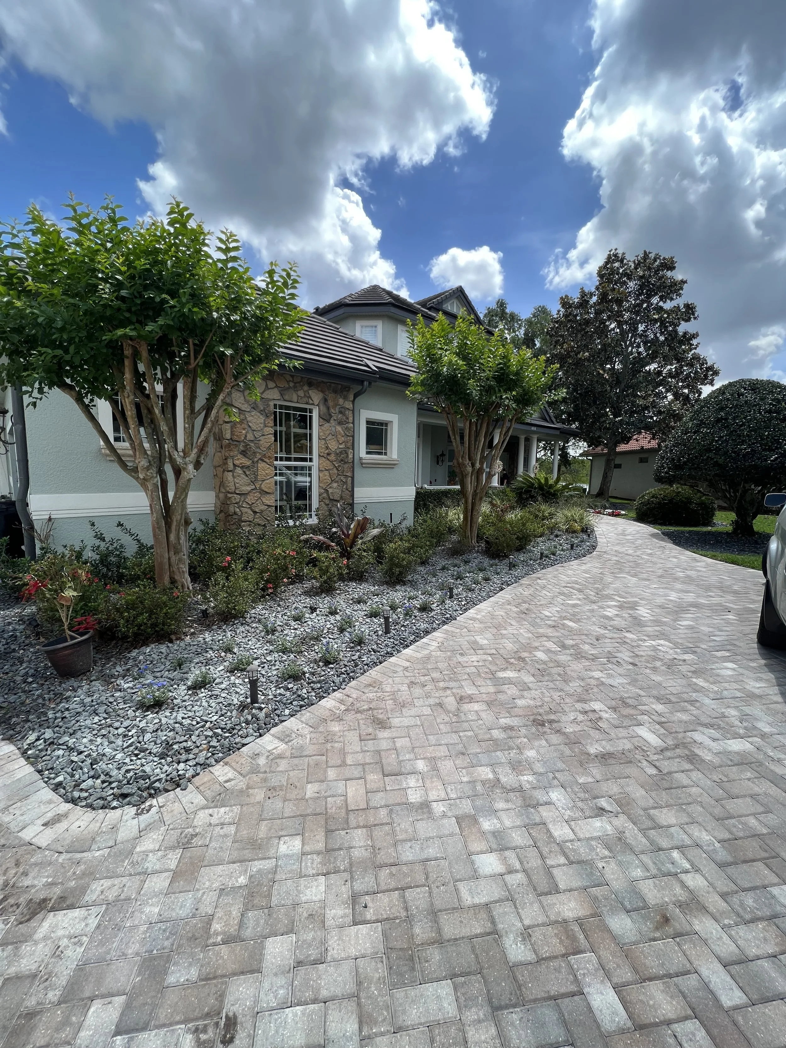 A landscaped front yard with a curved brick walkway, trees, shrubs, and a house with stone and siding exterior, under a partly cloudy sky.