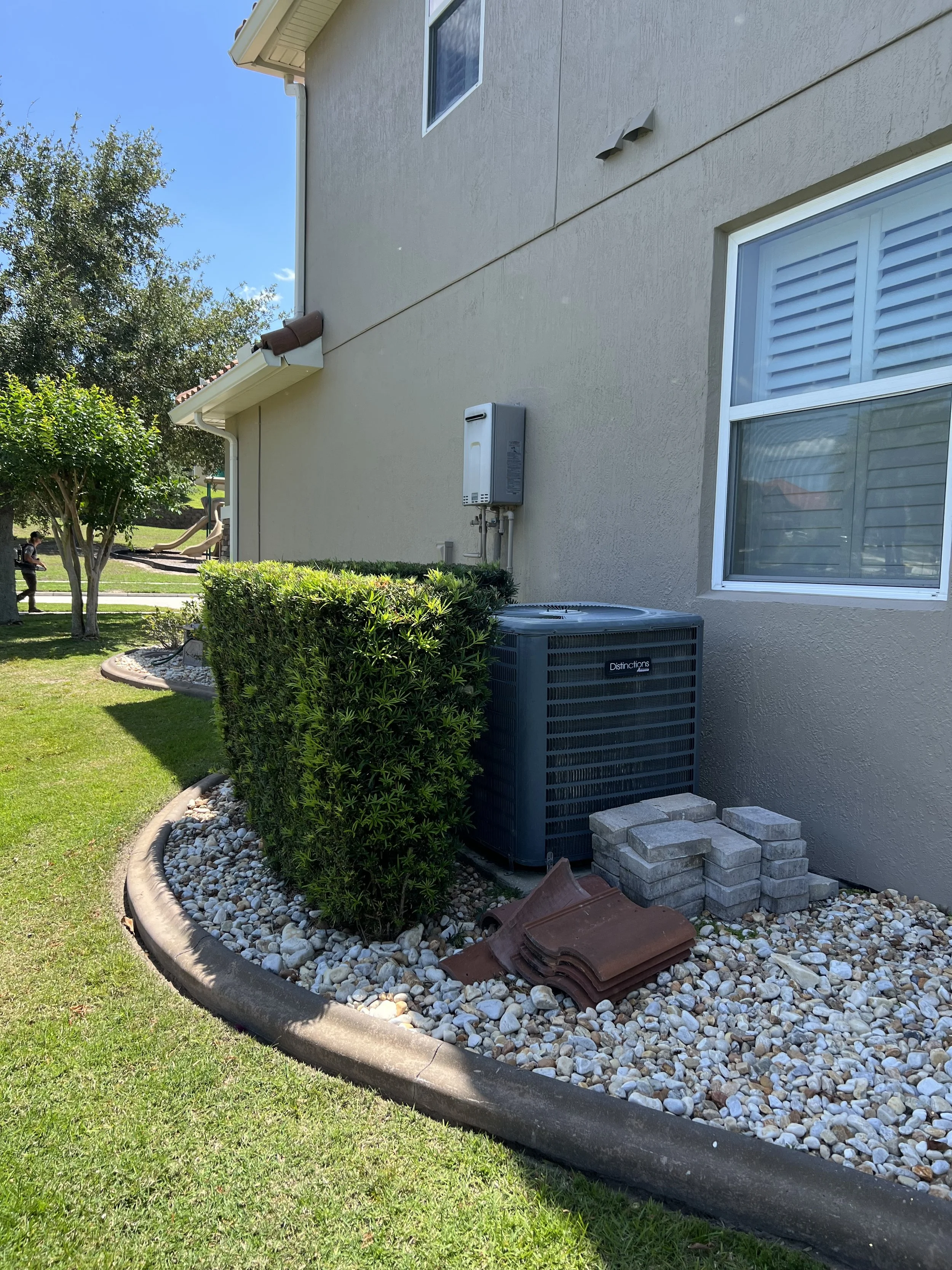 The side of a house with a beige wall, white-framed window with horizontal blinds, an air conditioning unit, a utility box, a stack of bricks, and a landscaped garden with rocks, bushes, and a tree. In the background, a person and a playground with s