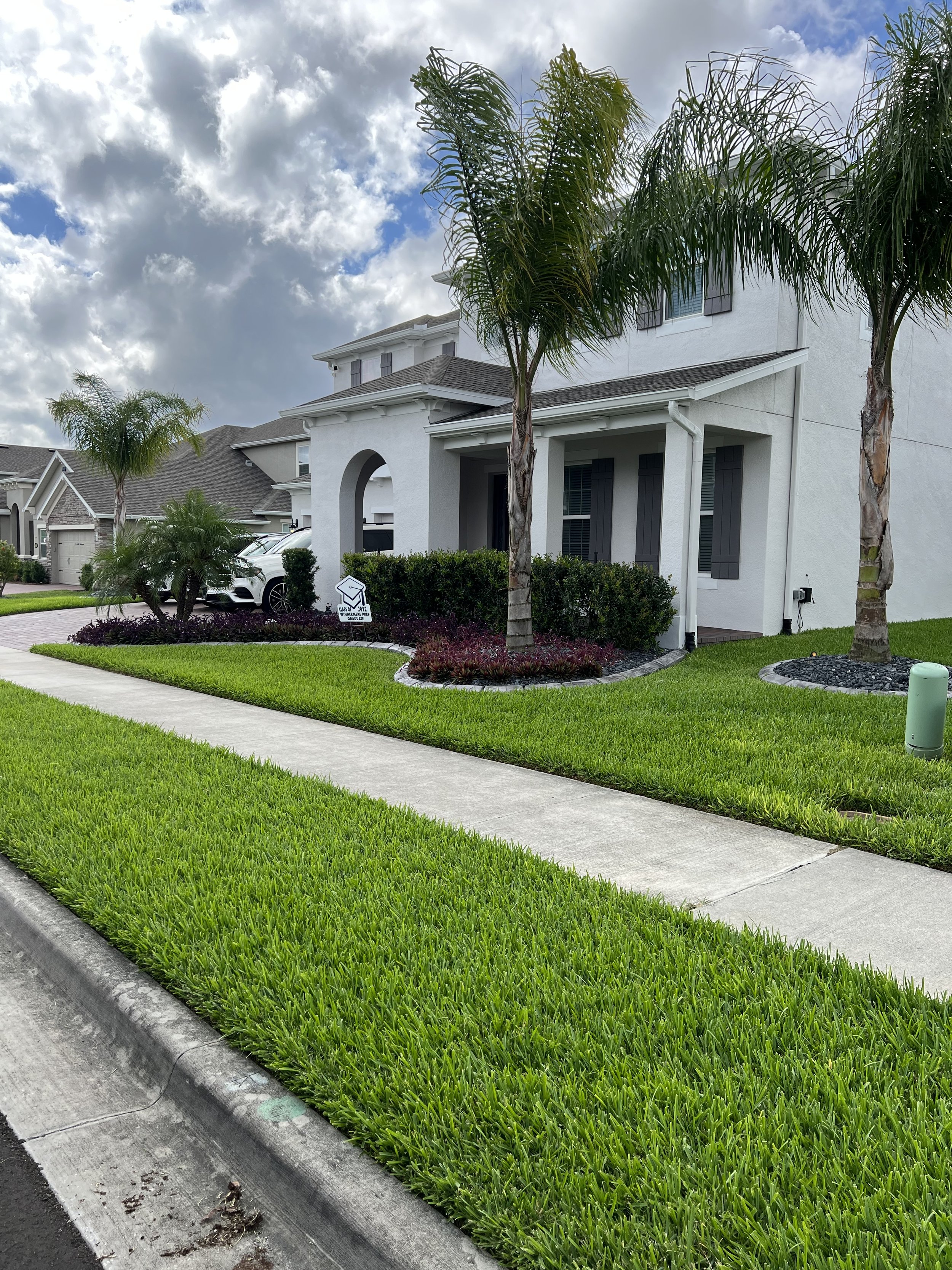 A suburban street with green grass, palm trees, mulch beds, and white houses with gray roofs under a partly cloudy sky.