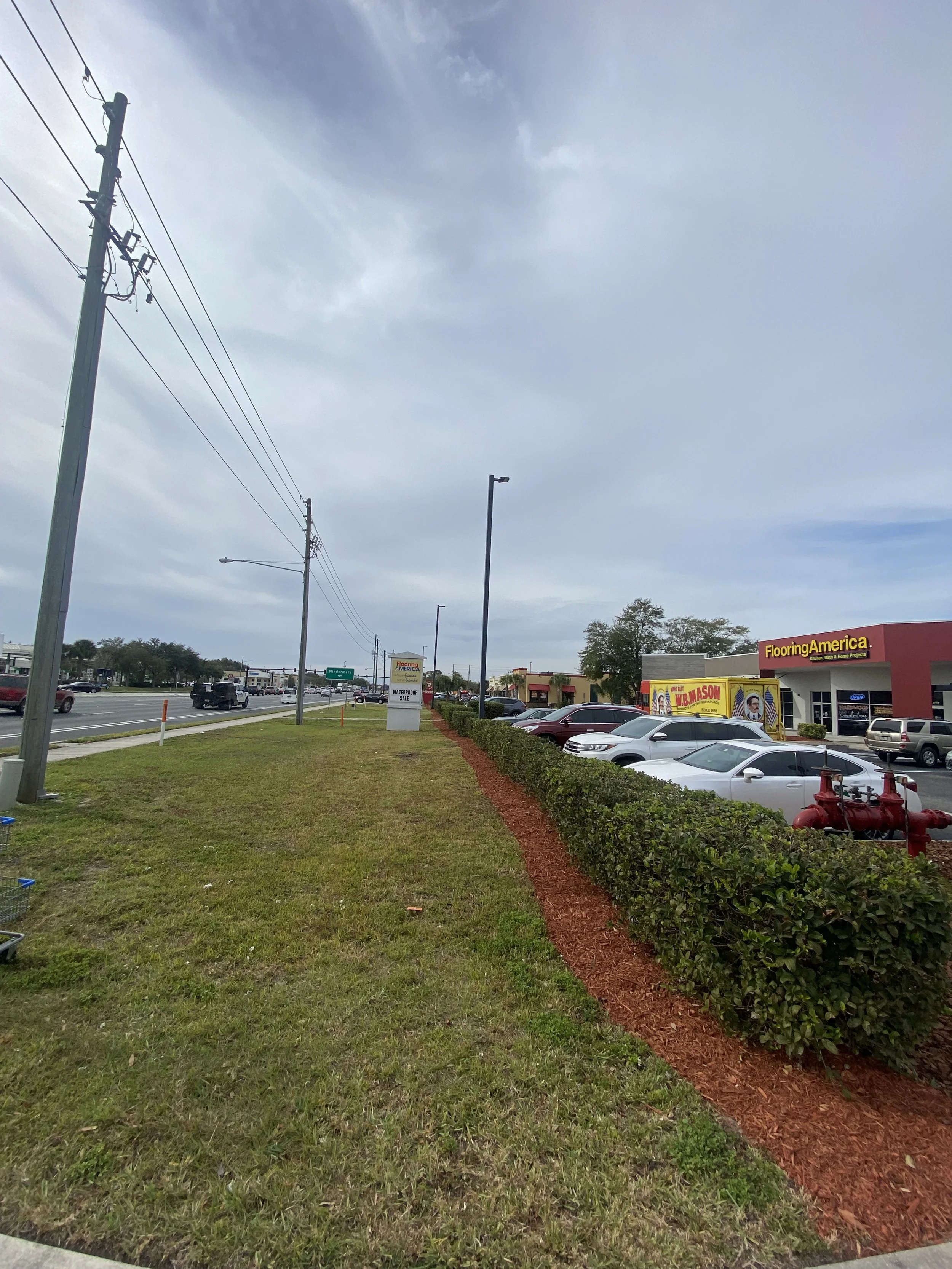 Parking lot adjacent to a strip mall under cloudy sky with utility pole, street lamps, and red mulch edging surrounding trimmed bushes.