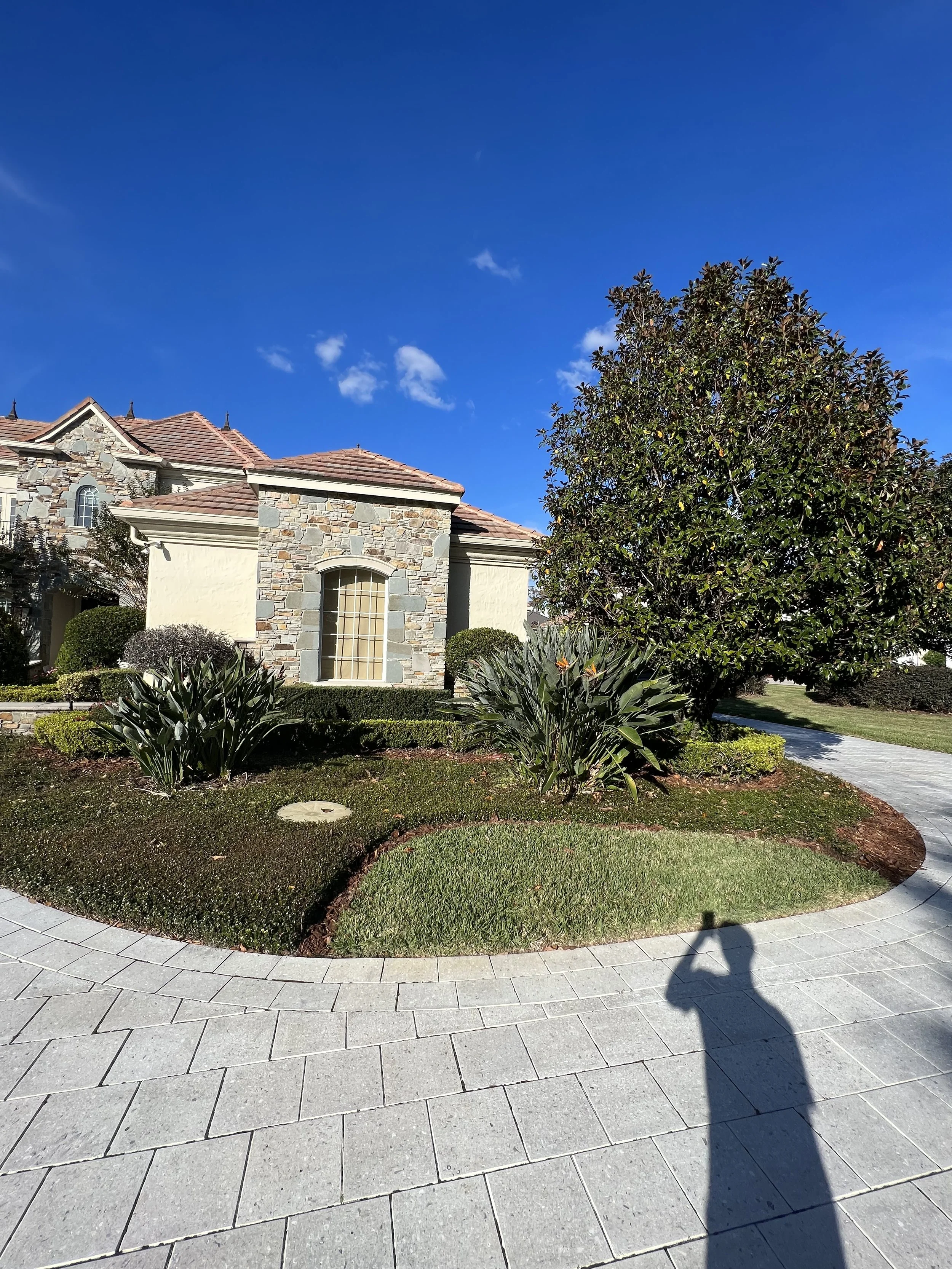 Front yard of a house with a stone facade, pink tile roof, well-maintained bushes, and a large tree. Paved walkway curves through the garden under a clear blue sky.
