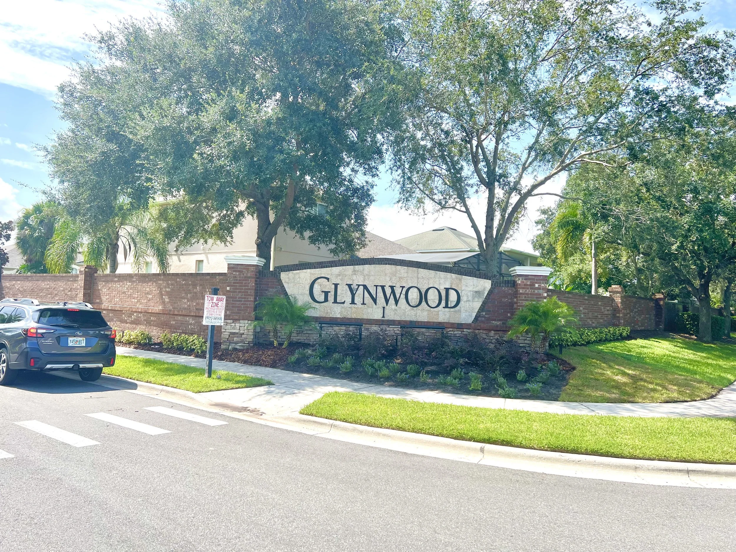 Large brick entrance sign with the word 'Glynnwood' in black letters, surrounded by trees and greenery, with parked cars in front and a street with a crosswalk.