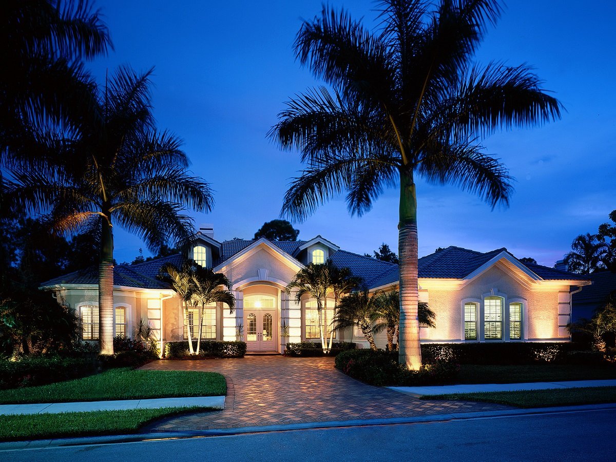 A large house with exterior lights at dusk, surrounded by palm trees and a neatly maintained lawn, with a paved walkway leading to the front door.