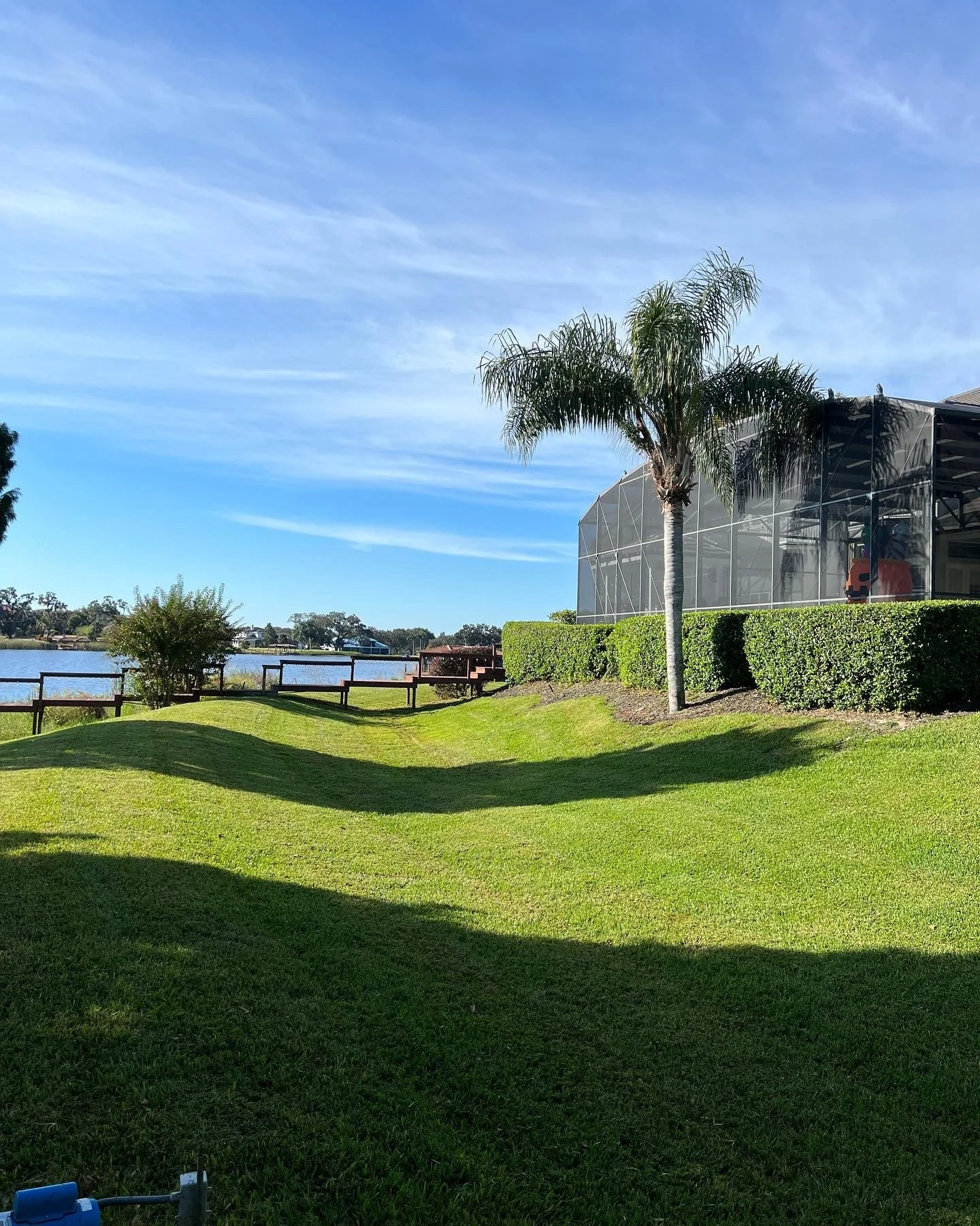 A lush green lawn near a body of water with a wooden railing, a bush, and a tall palm tree, with a building with a screened porch and a cloudy blue sky in the background.