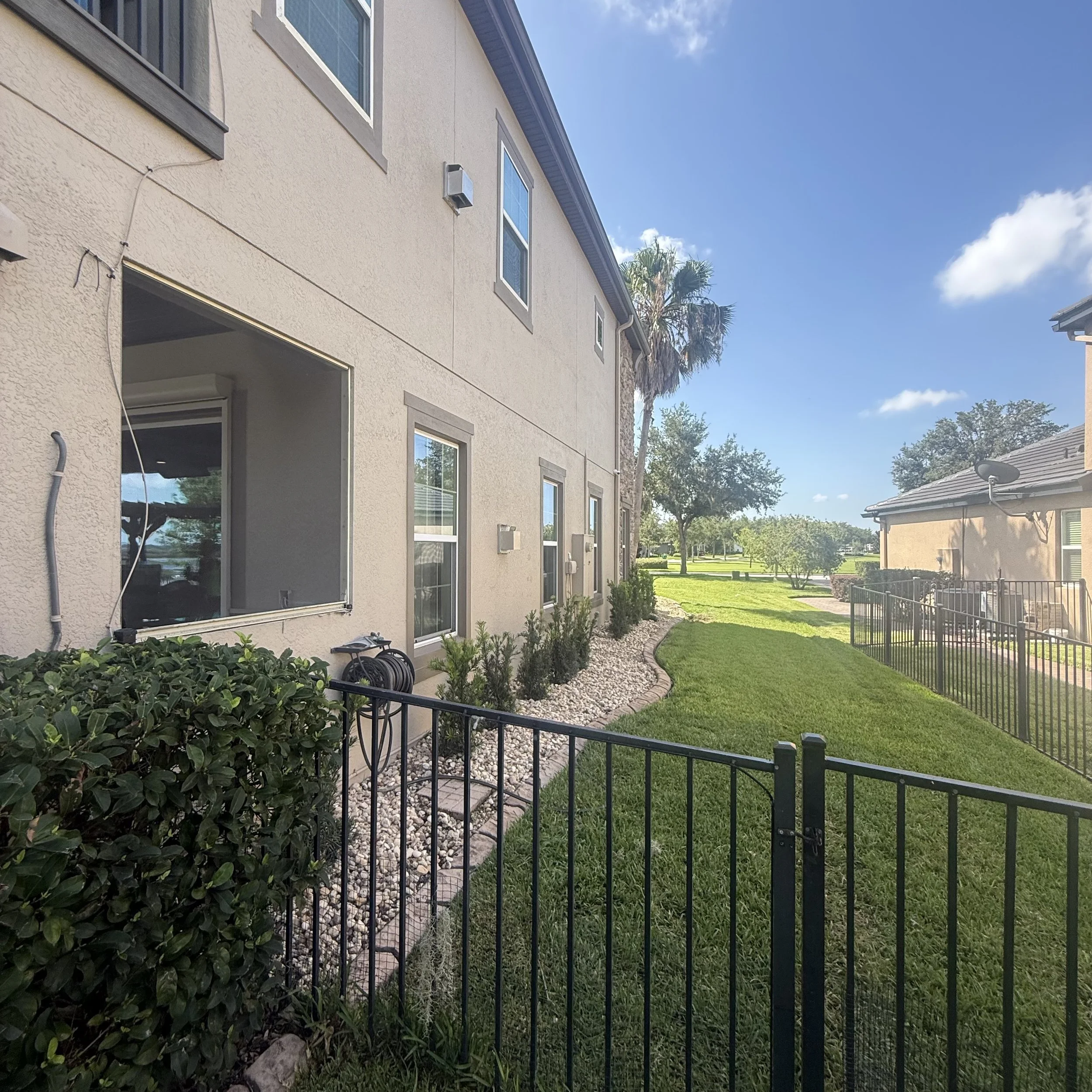 View of the exterior of a beige residential building with several windows, some open, and a small balcony. The backyard features a neatly maintained lawn, a garden bed with bushes and plants, a rock mulch border, and a black metal fence surrounding t