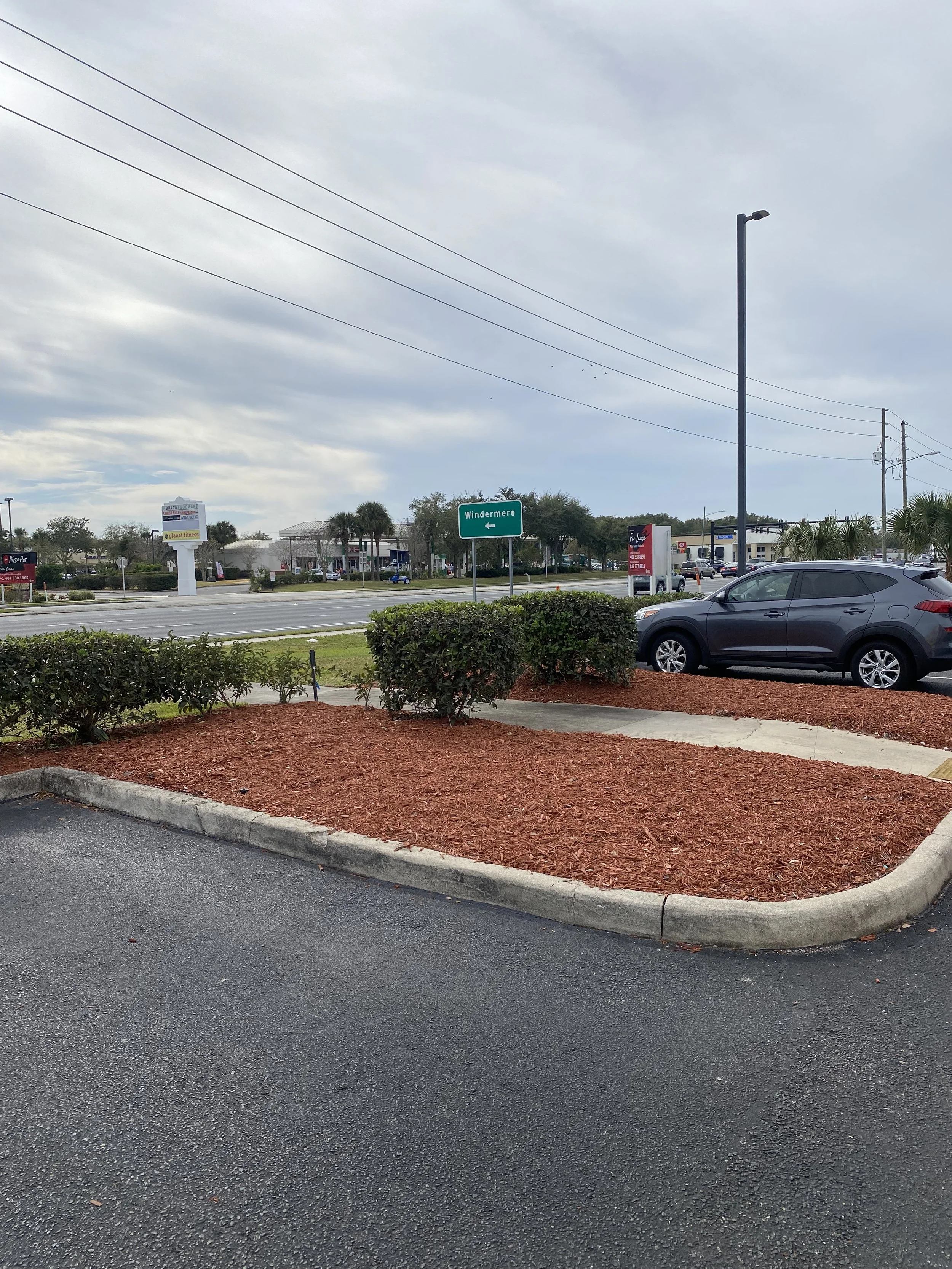 A parking lot with a landscaped island covered in red mulch, bushes, and a small concrete border. In the background, there are commercial buildings, a street sign pointing left to Windermere, and parked cars.