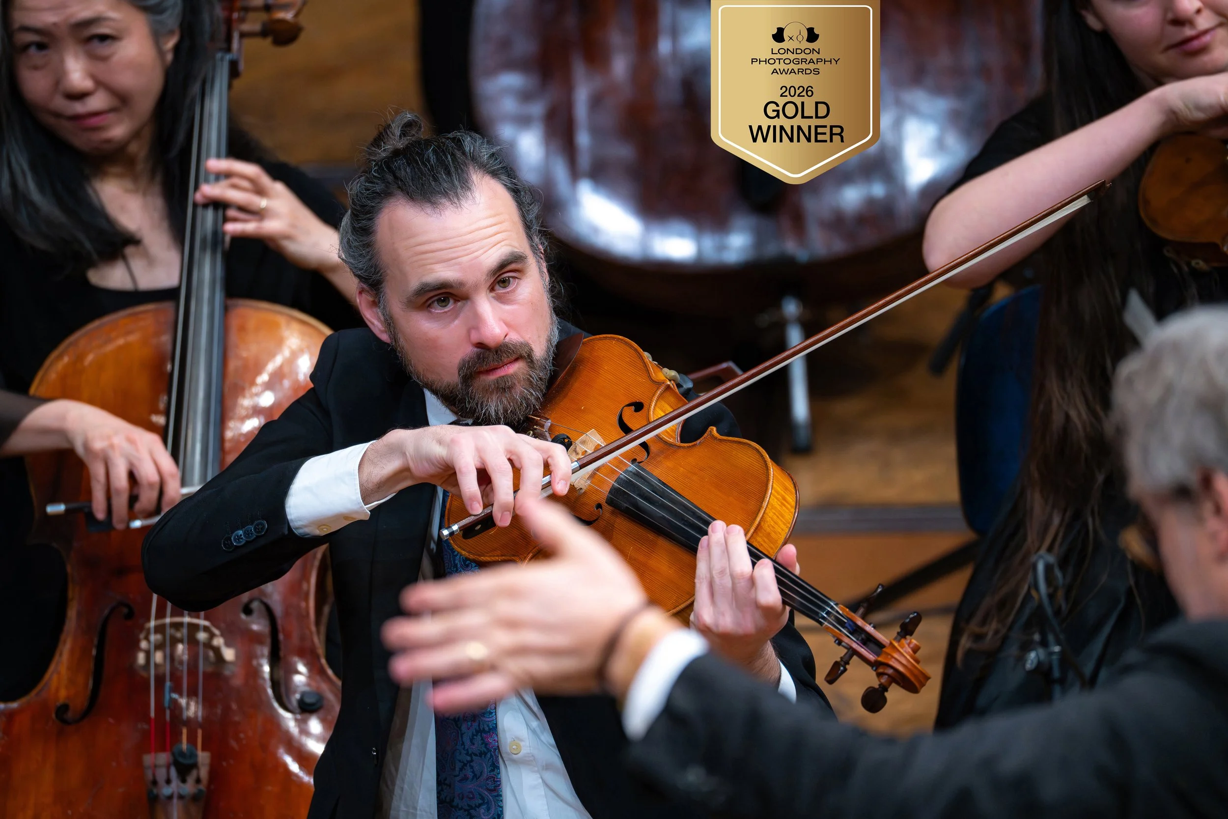 Ein Mann spielt Geige während eines Orchesters, umgeben von anderen Musikern, in einem Konzertsaal. Es gibt eine Auszeichnung für den Goldgewinner bei den London Photography Awards 2026 in der oberen rechten Ecke.
