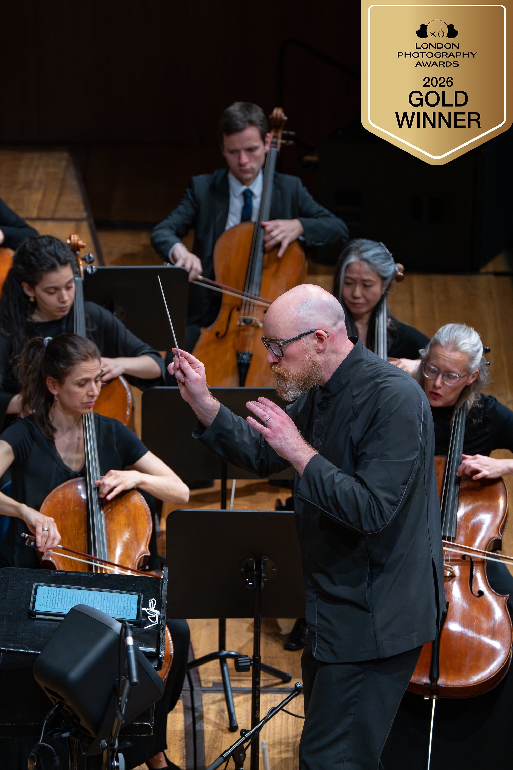 Ein Orchesterproben mit mehreren Musikern, die Violinen und ein Cello spielen, und einem Dirigenten, der dirigiert. Im oberen rechten Bereich befindet sich eine Goldmedaille mit der Aufschrift 'London Photography Awards 2026 Gold Winner'.