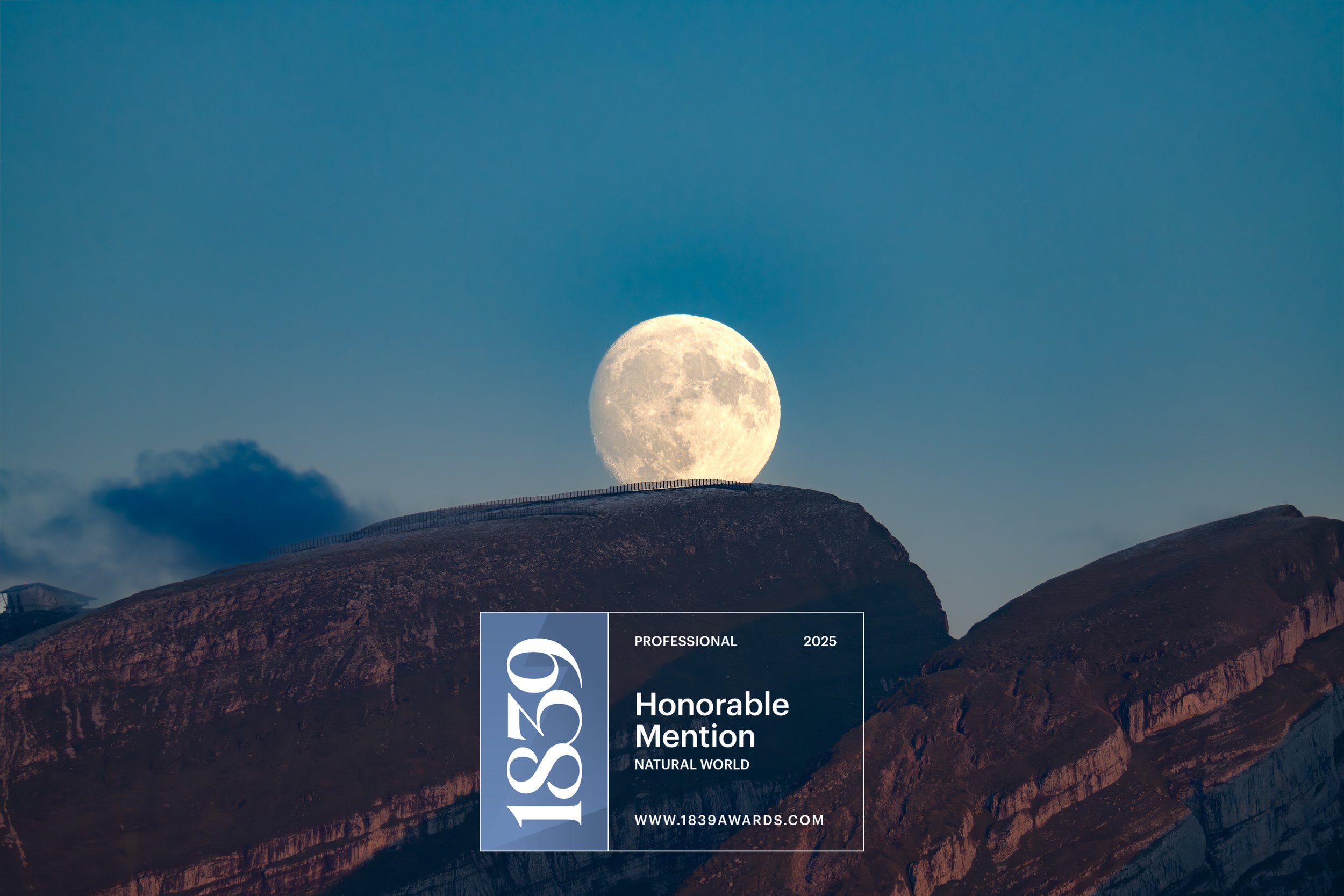 Der Mond steht hoch am Himmel über einer Berglandschaft, im Hintergrund Wolken, vorne ein Hügel mit einem Zaun. Über dem Bild liegt ein Zertifikat mit dem Text "Honourable Mention" und Informationen zu den 1839 Awards.