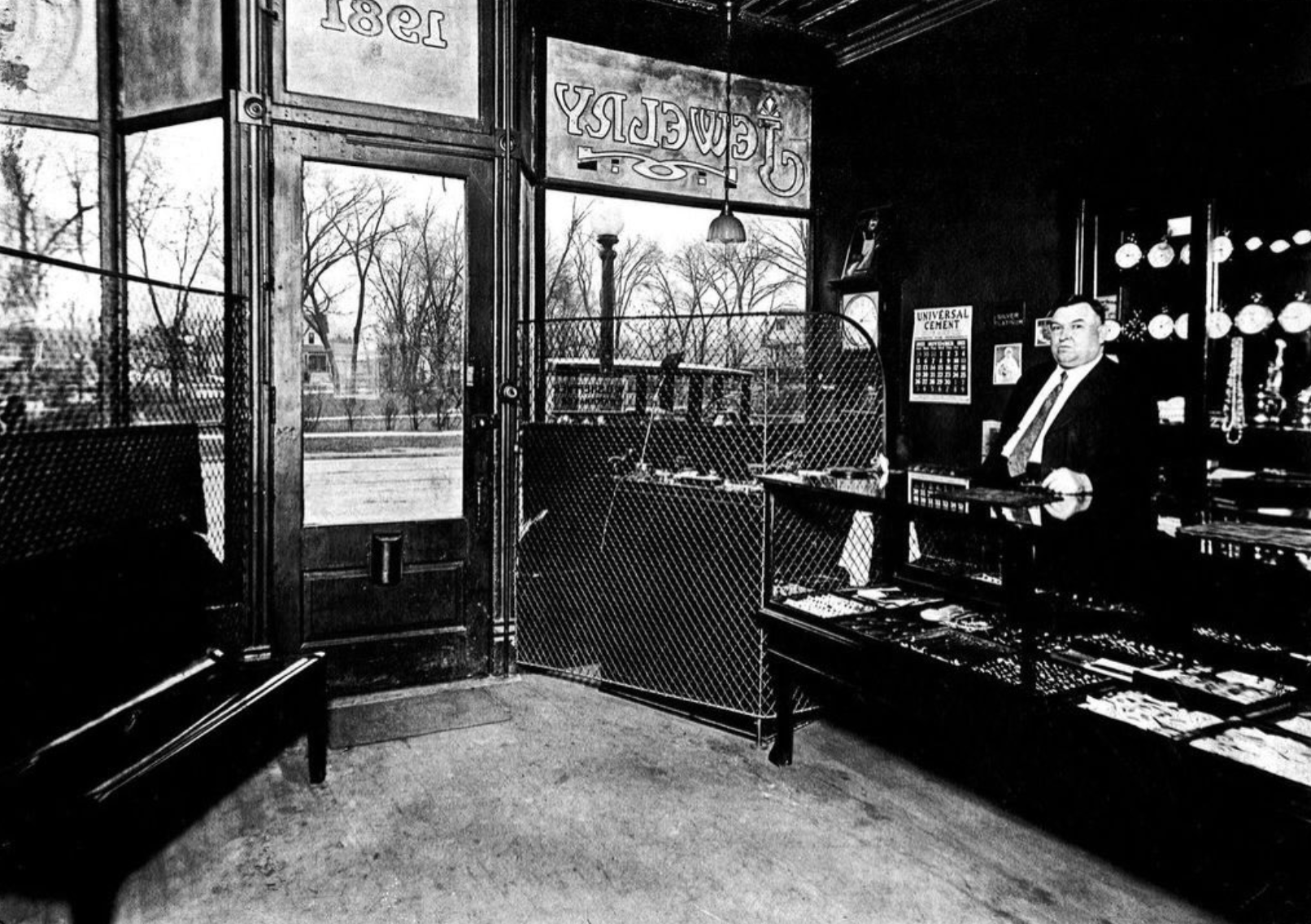 Black and white photo of the interior of a vintage shop, featuring a man standing behind a display counter with various items, a large open door with windows showing trees outside, signs that read 'Universal Cement' and 'Groceries', and a shelf with lighting fixtures in the background.