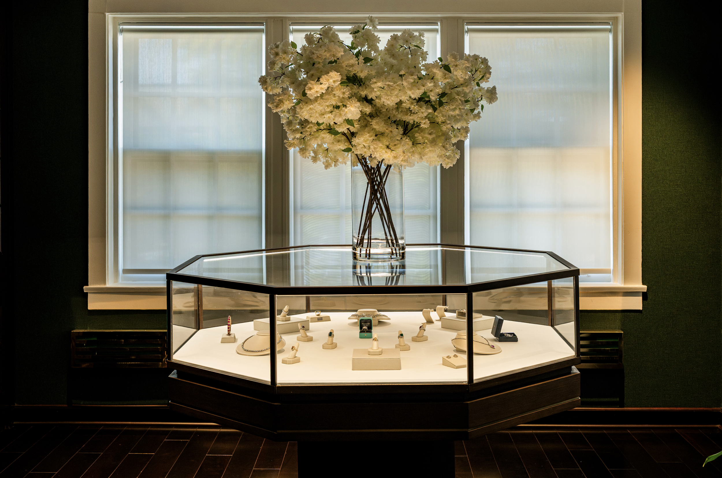 A jewelry display case with various jewelry pieces, a large vase of white flowers on top, and a window with closed blinds behind it.