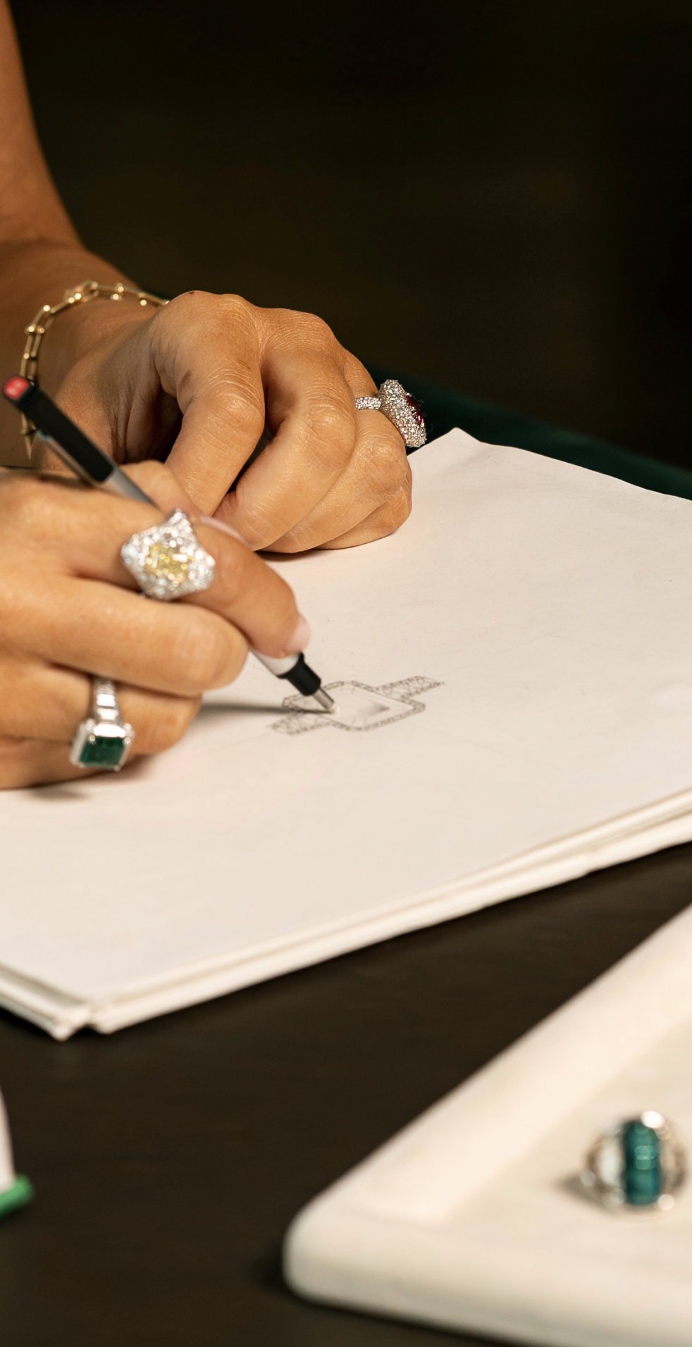 Close-up of a person's hands drawing a sketch of a square with a smaller square inside on a white paper using a black pen. The person is wearing multiple rings, including a large diamond ring, rings with colorful stones, a bracelet, and a large ring with a green stone. The background is dark, and additional jewelry is visible on the table.