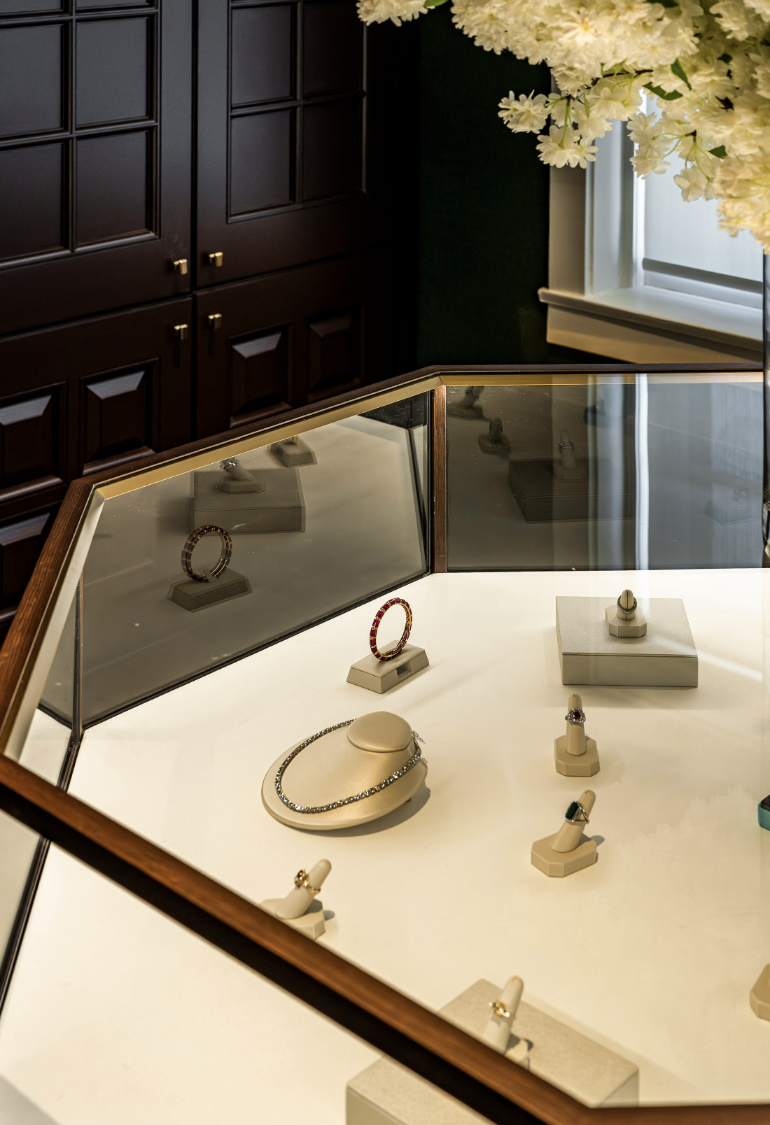 Jewelry display case showcasing earrings, rings, and necklaces in a boutique with dark cabinets and a window with white flowers.