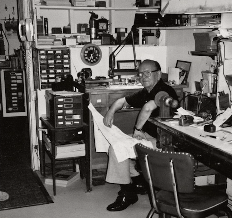 A man wearing glasses sitting at a cluttered workbench in a workshop or office, surrounded by tools, papers, and shelves filled with small drawers and books.
