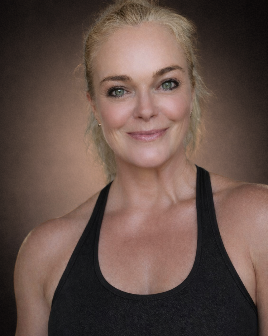 Portrait of a smiling woman with blonde hair and blue eyes, wearing a black sports top against a dark background.