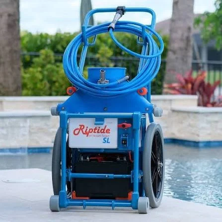 Blue portable pool vacuum with wheels, hoses, and a handle, labeled Riptide SL, on a poolside.