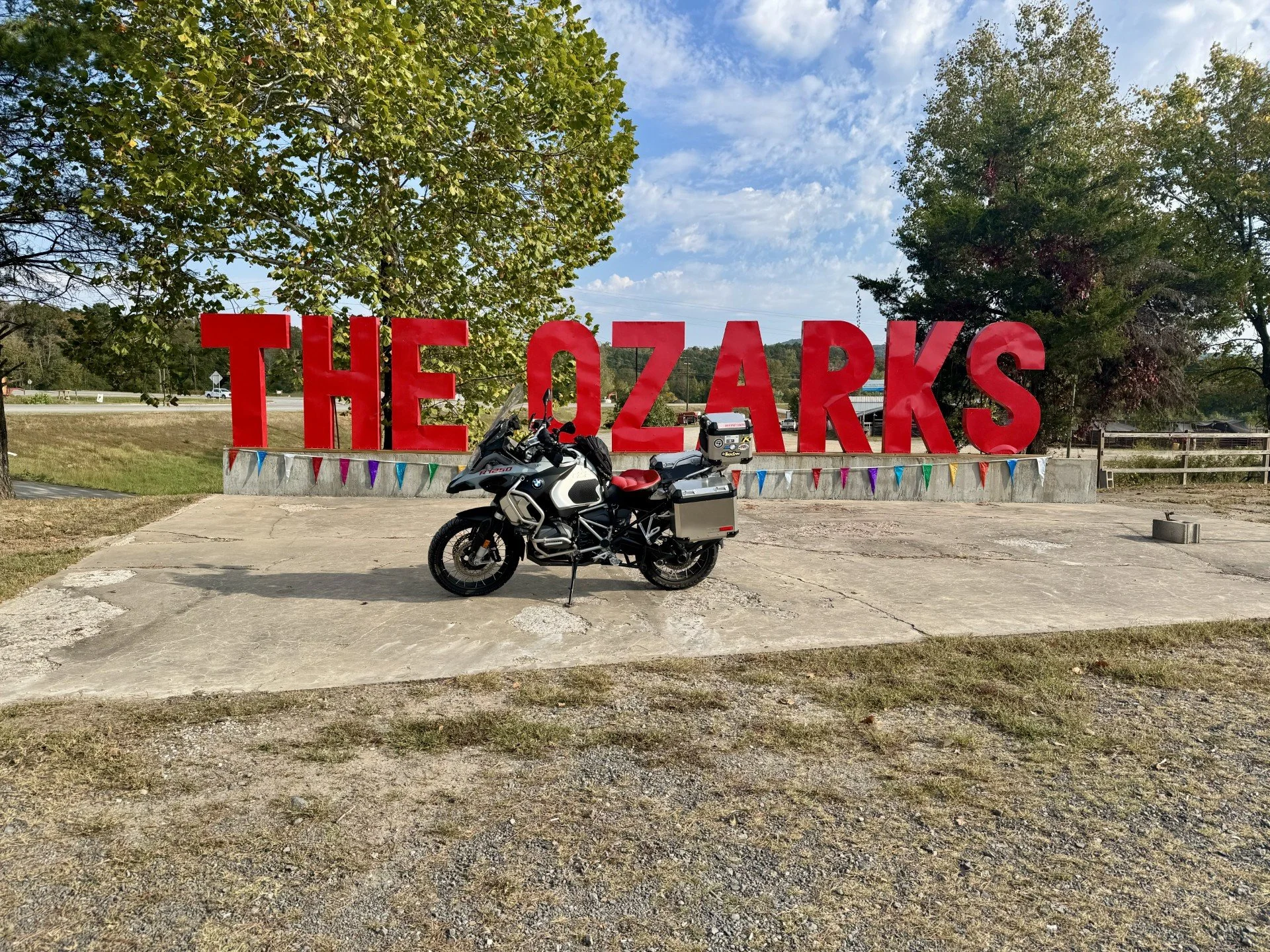 A large red sign spelling 'THE OZARKS' on a concrete platform outside, with a black and white motorcycle parked in front. Trees and a partly cloudy sky in the background.